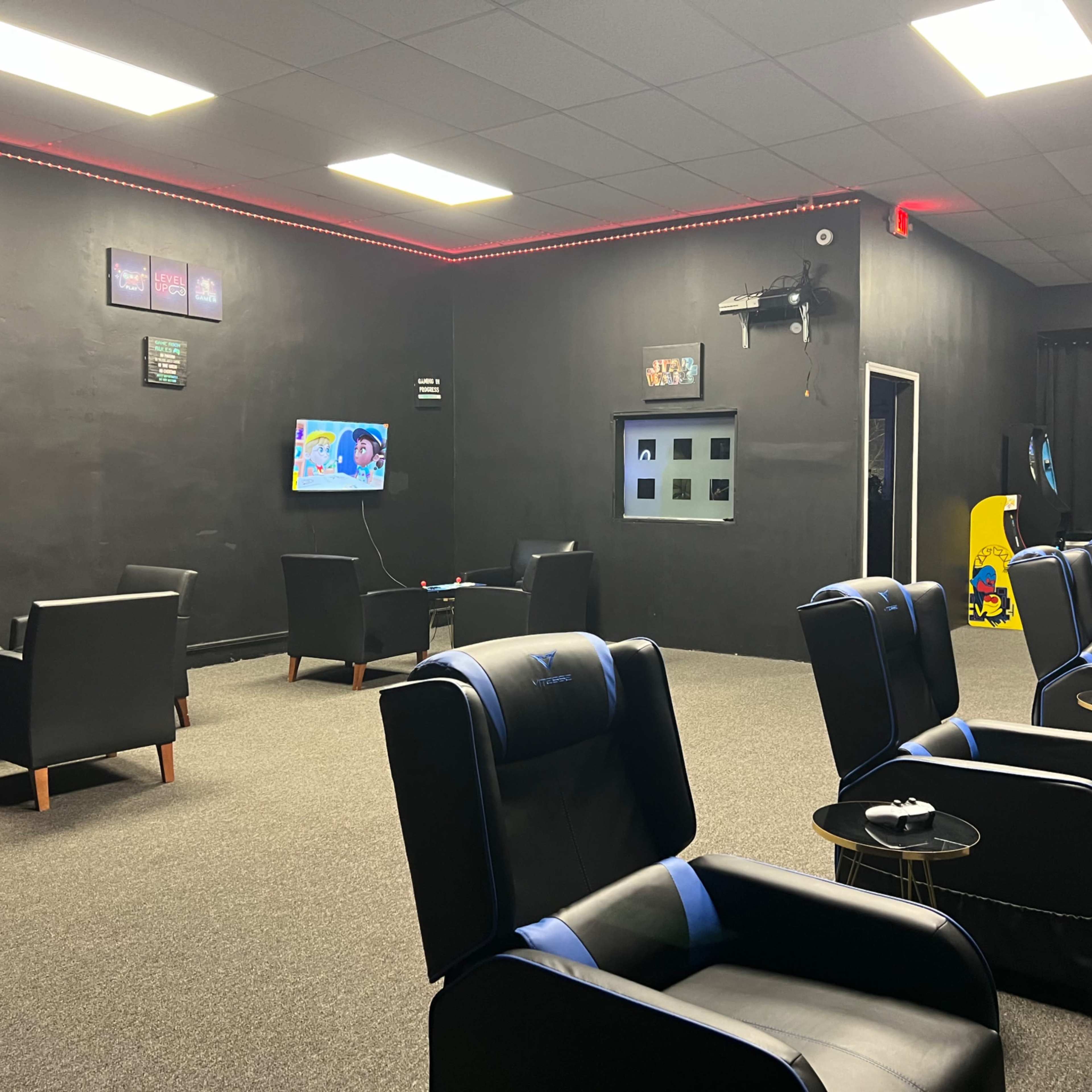 A dimly lit lounge area with black walls, featuring multiple seating chairs arranged in rows facing televisions.