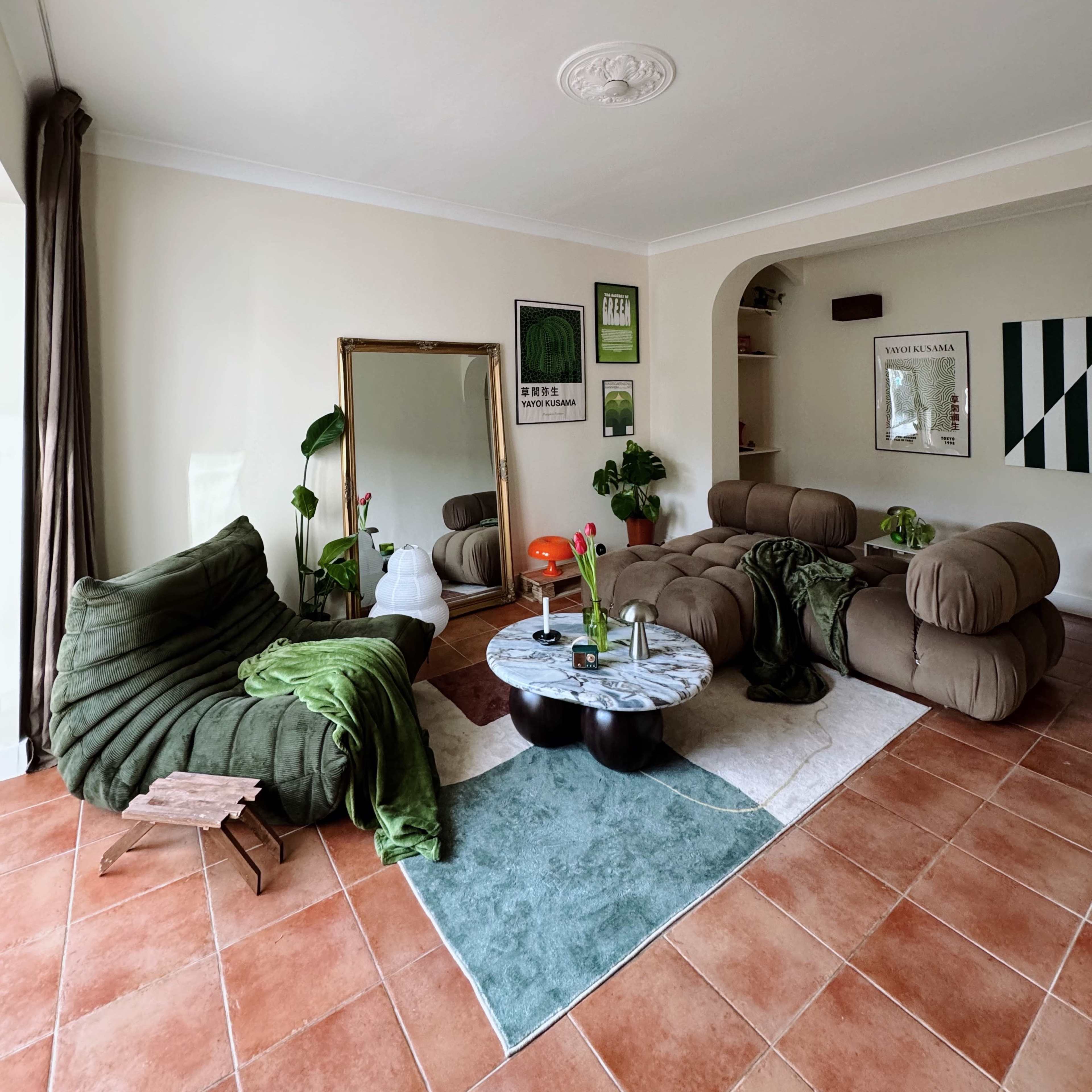 The living room features two modern sofas, a round marble coffee table, and plants, arranged on a patterned area rug with terracotta tile flooring.