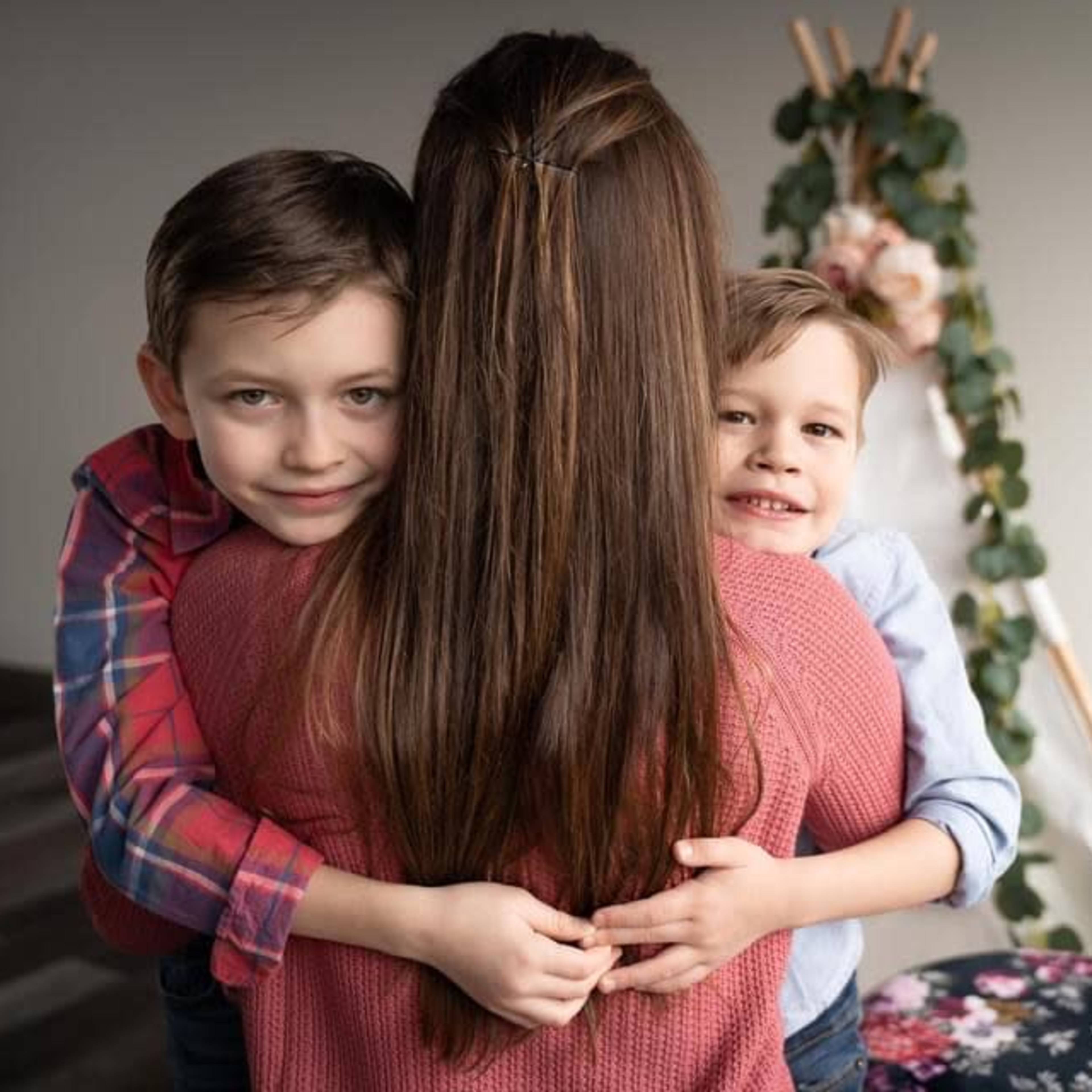A woman stands with her two children, who are hugging her from behind, in a room decorated with flowers and greenery.