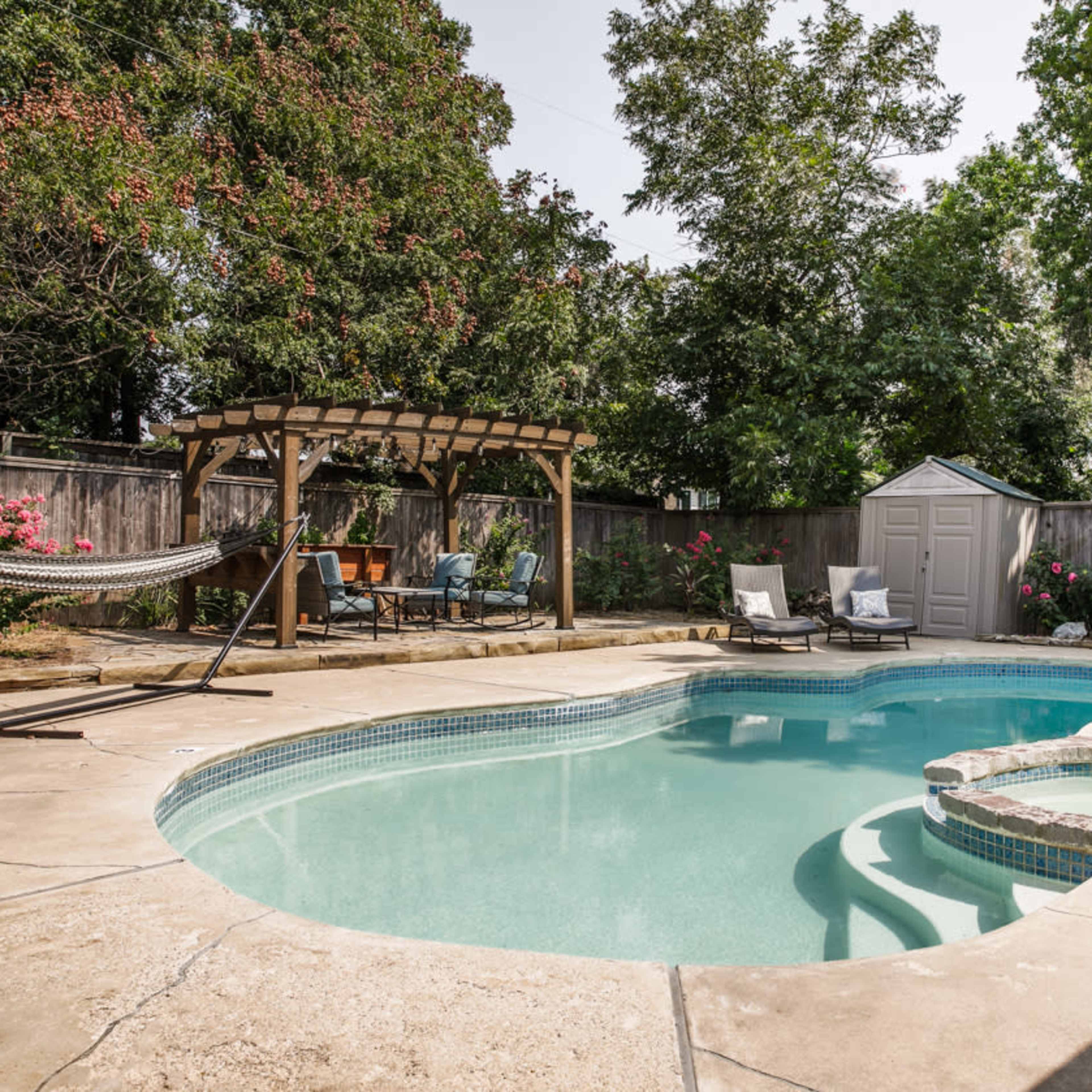 A landscaped backyard features a swimming pool with a built-in spa, surrounded by a wooden pergola, hammock, and seating area.
