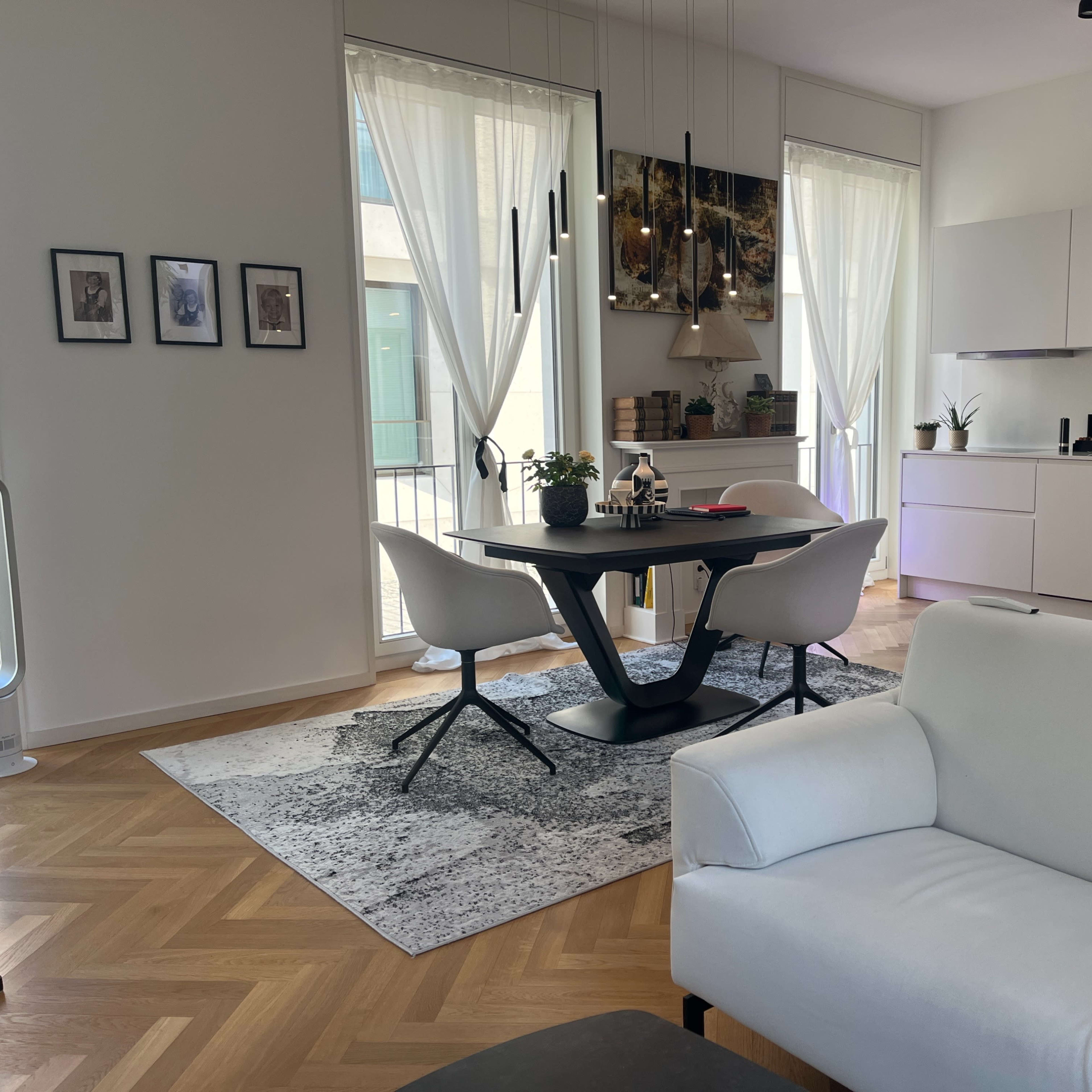A modern living space featuring a dining area with a round black table and white chairs, a white sofa in the foreground, and a kitchenette with sleek cabinetry in the background.