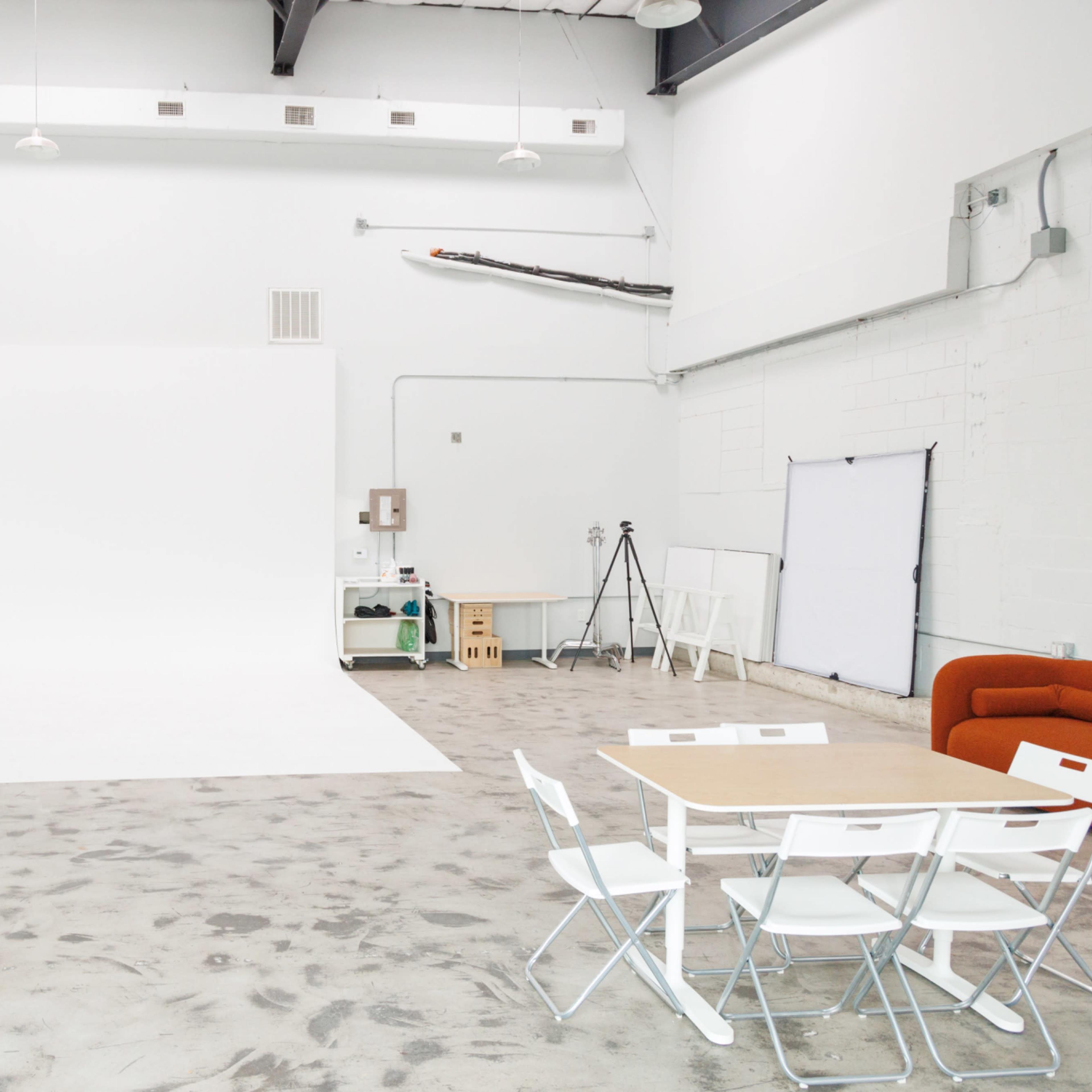 A spacious white studio with a clean backdrop, a table with chairs, and various equipment arranged in the corners.