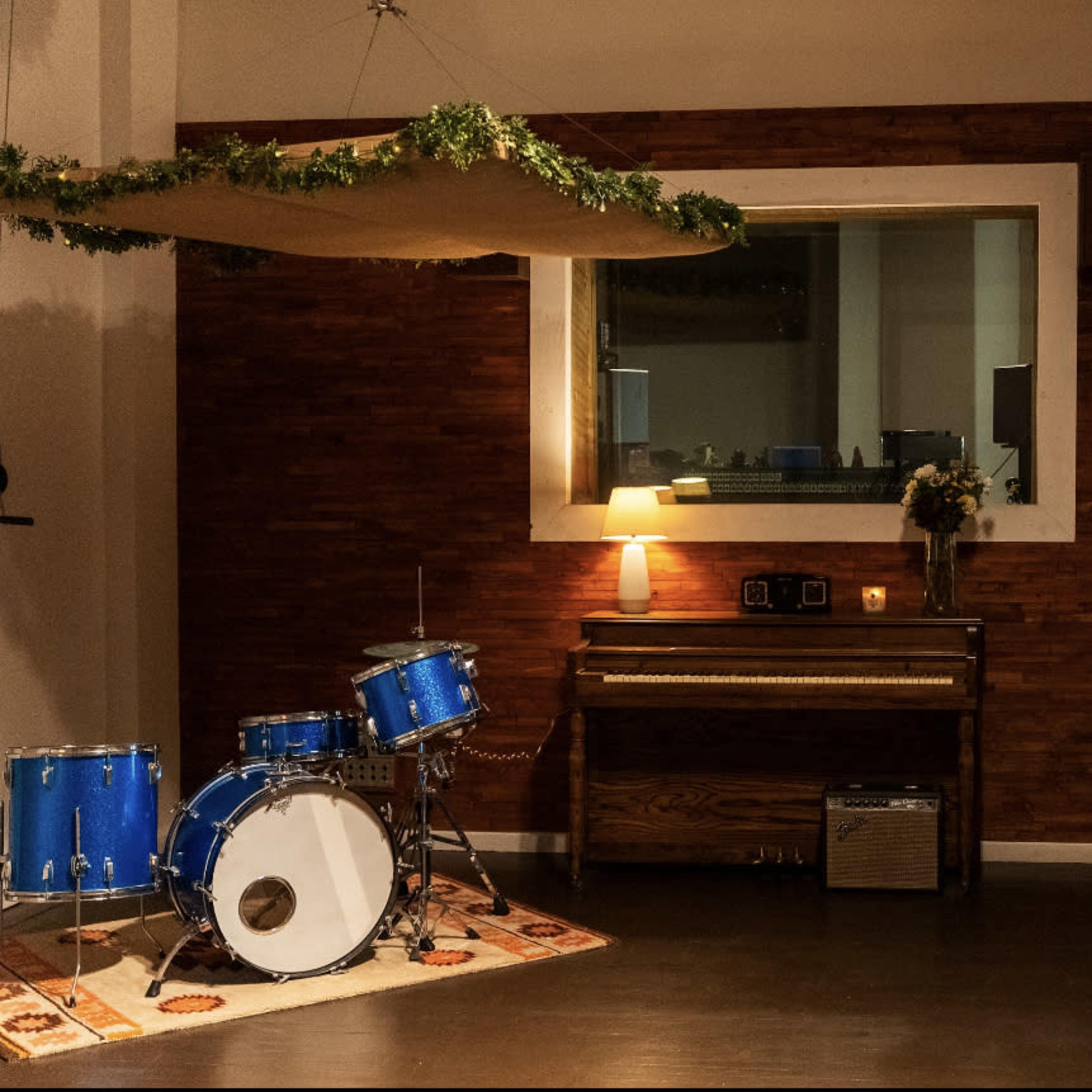 The scene features a music room with blue drums, a piano, and an amplifier, set against a wooden wall and illuminated by a lamp.