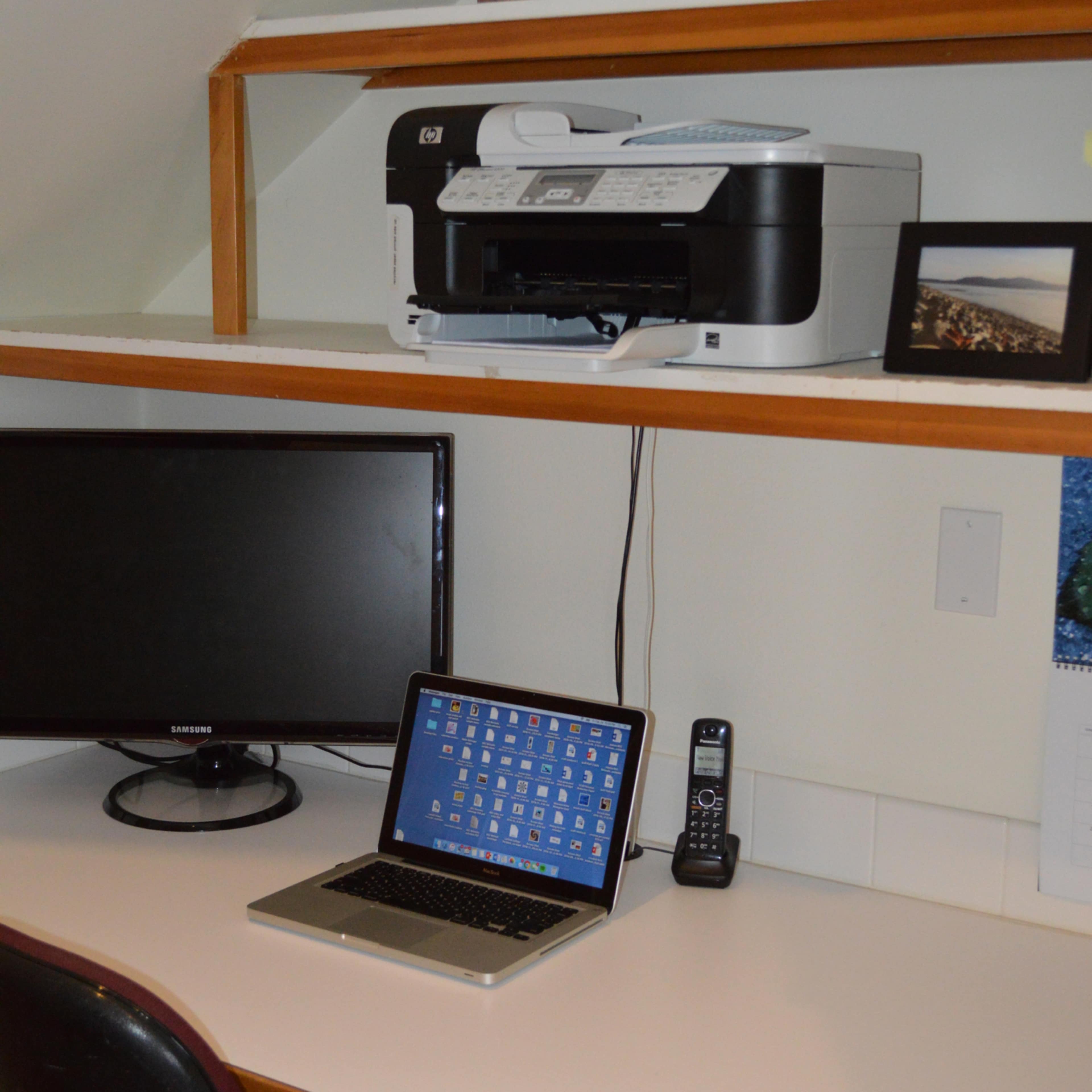 A home office setup featuring a printer on a shelf, a computer monitor and a laptop on a desk, along with a telephone and a framed photo.