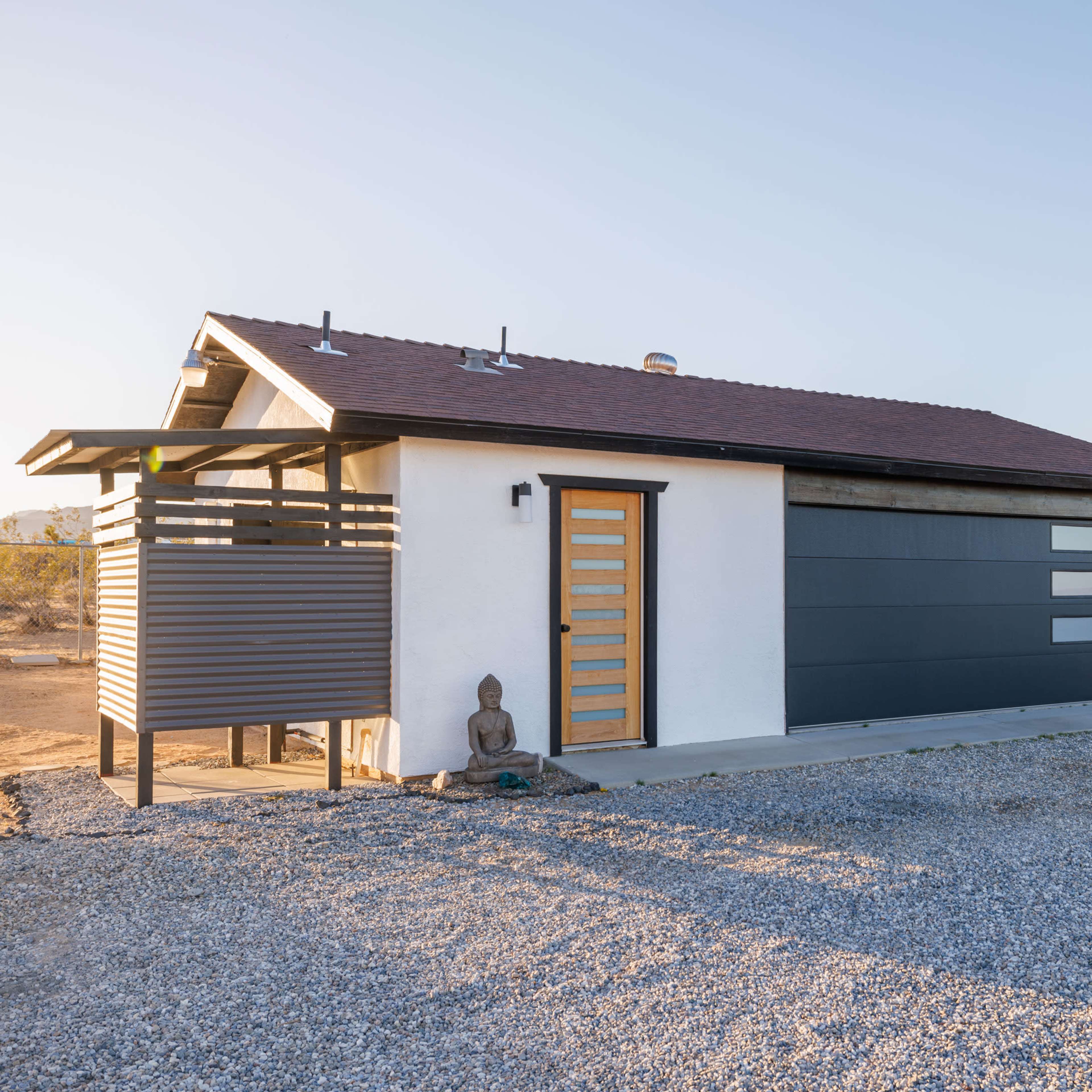 A modern single-story house features a gray and white exterior with a distinctive front door and a gravel driveway.