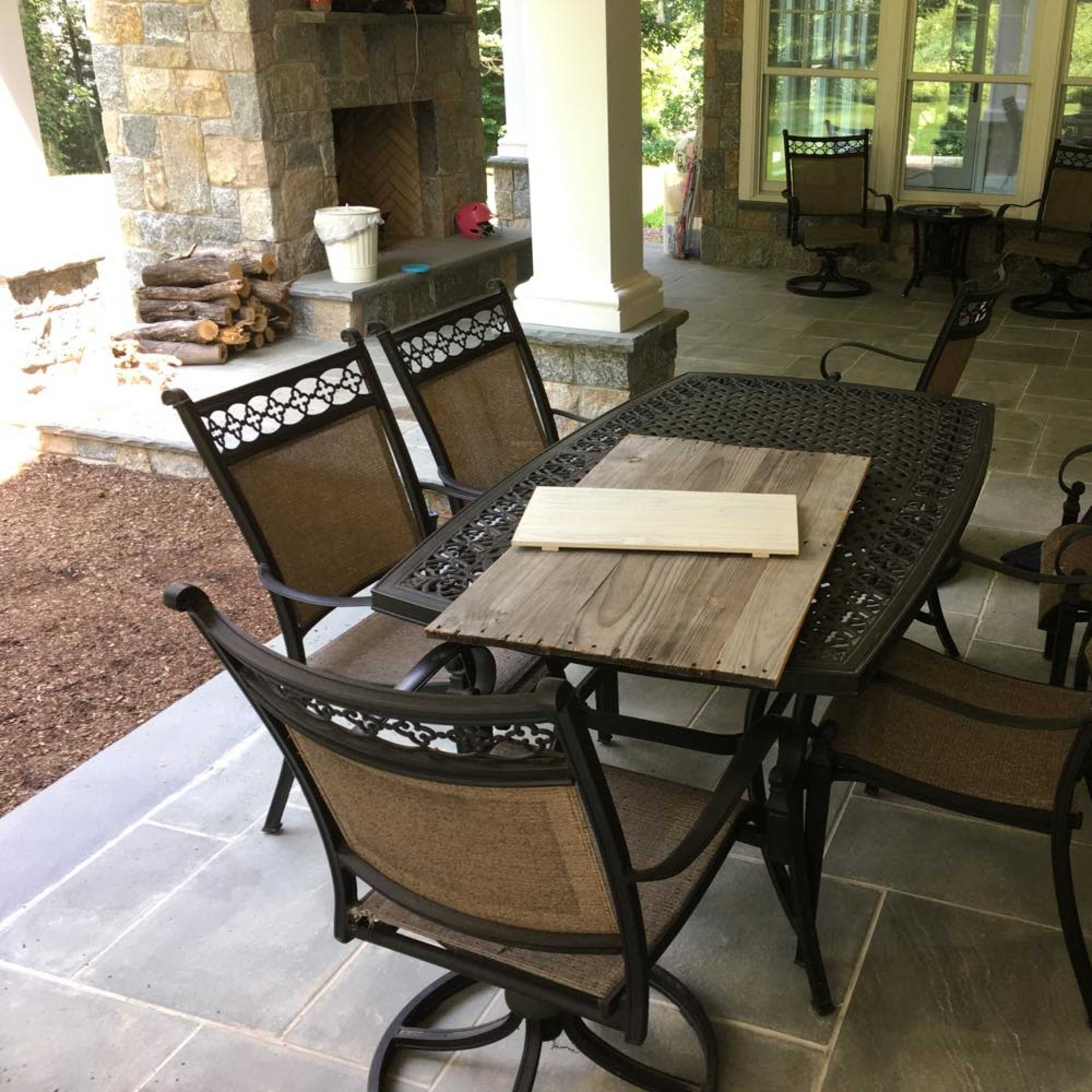 A patio area features a metal dining table surrounded by several chairs, with a wooden board placed on top of the table.