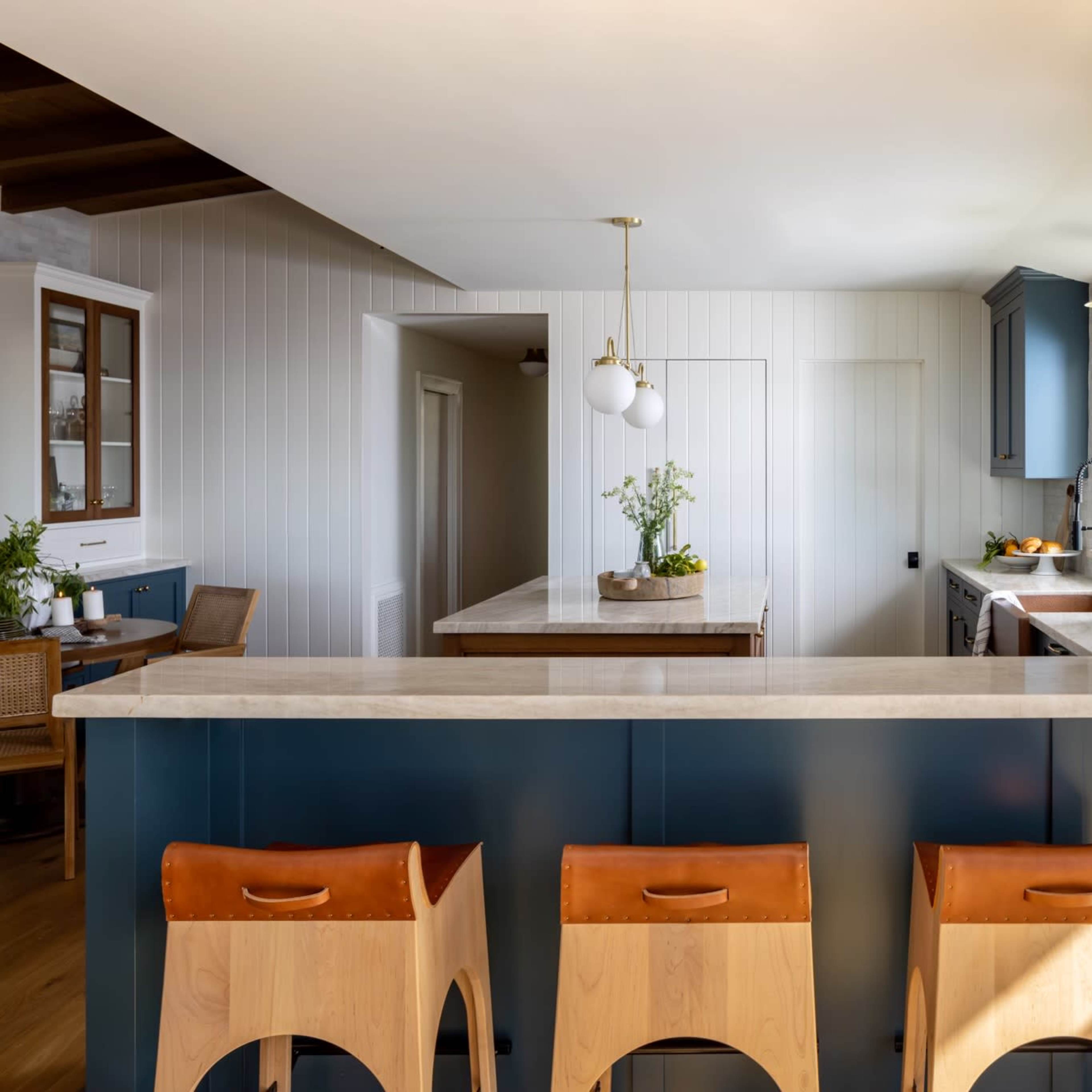 A modern kitchen features a large island with a light-colored countertop and three wooden stools, surrounded by navy blue cabinets and a bright, airy space.