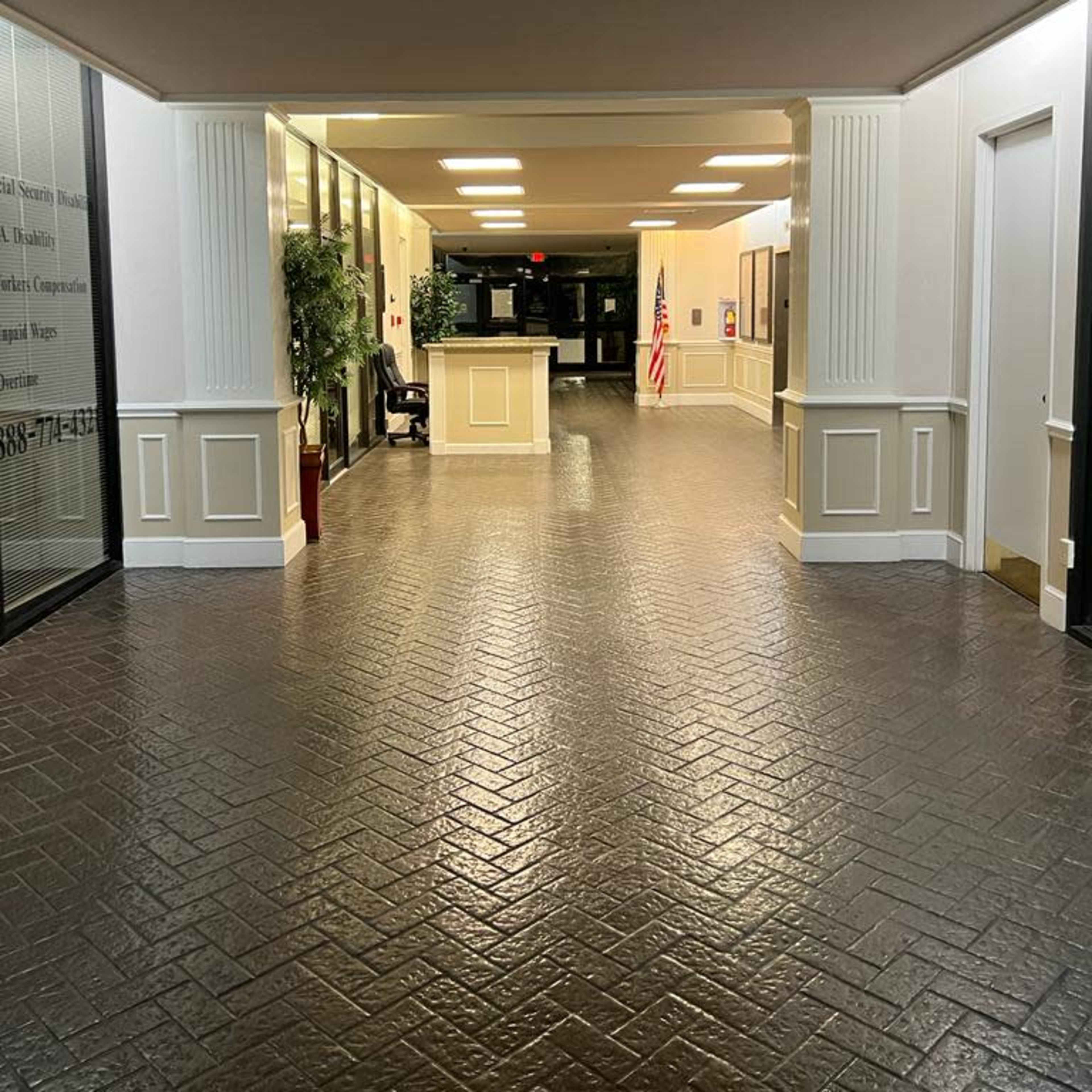 A corridor with textured flooring, lights overhead, and a reception area at the end, featuring an American flag and potted plants.