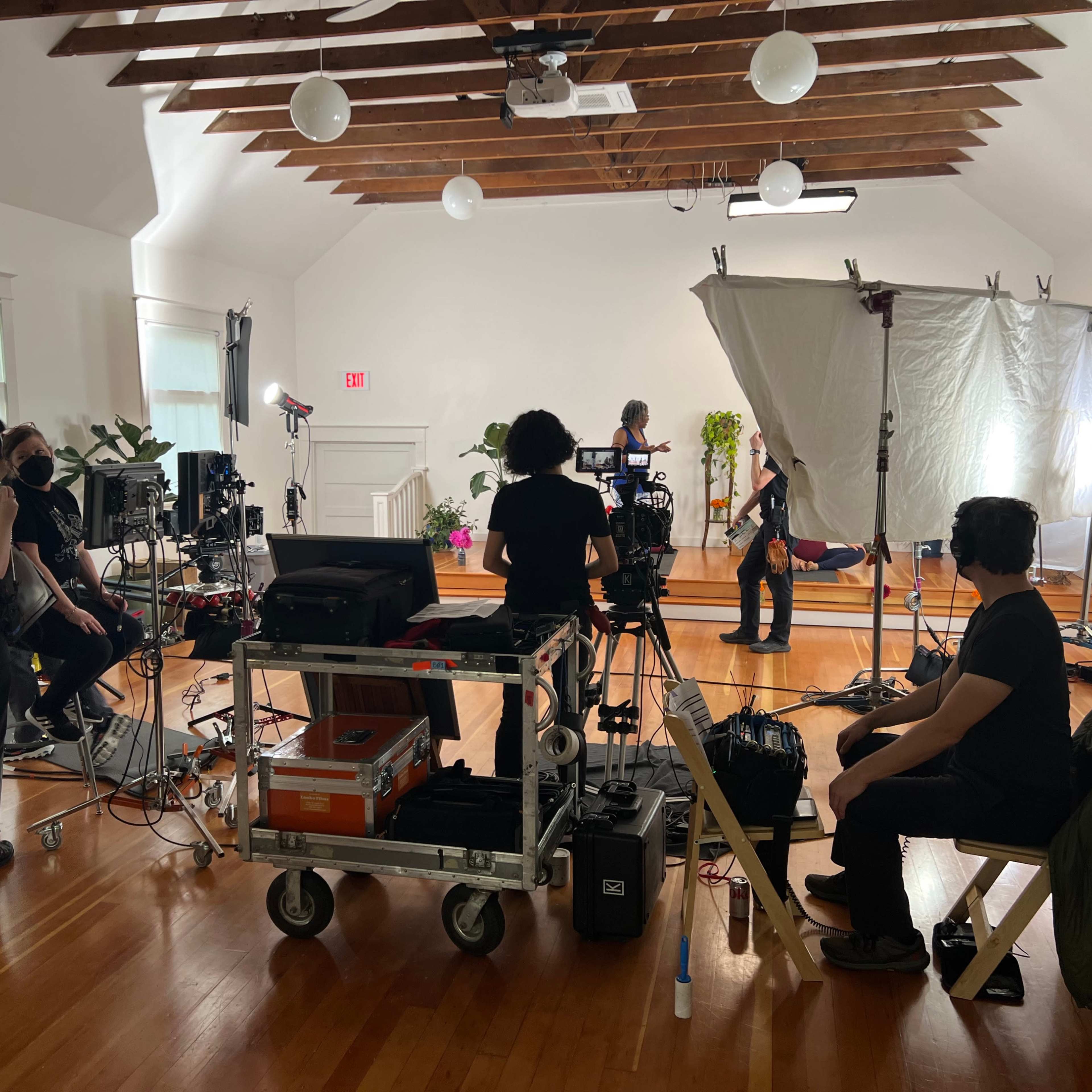 A film crew is set up inside a well-lit room, working with lights, cameras, and a large reflector in preparation for a shoot.