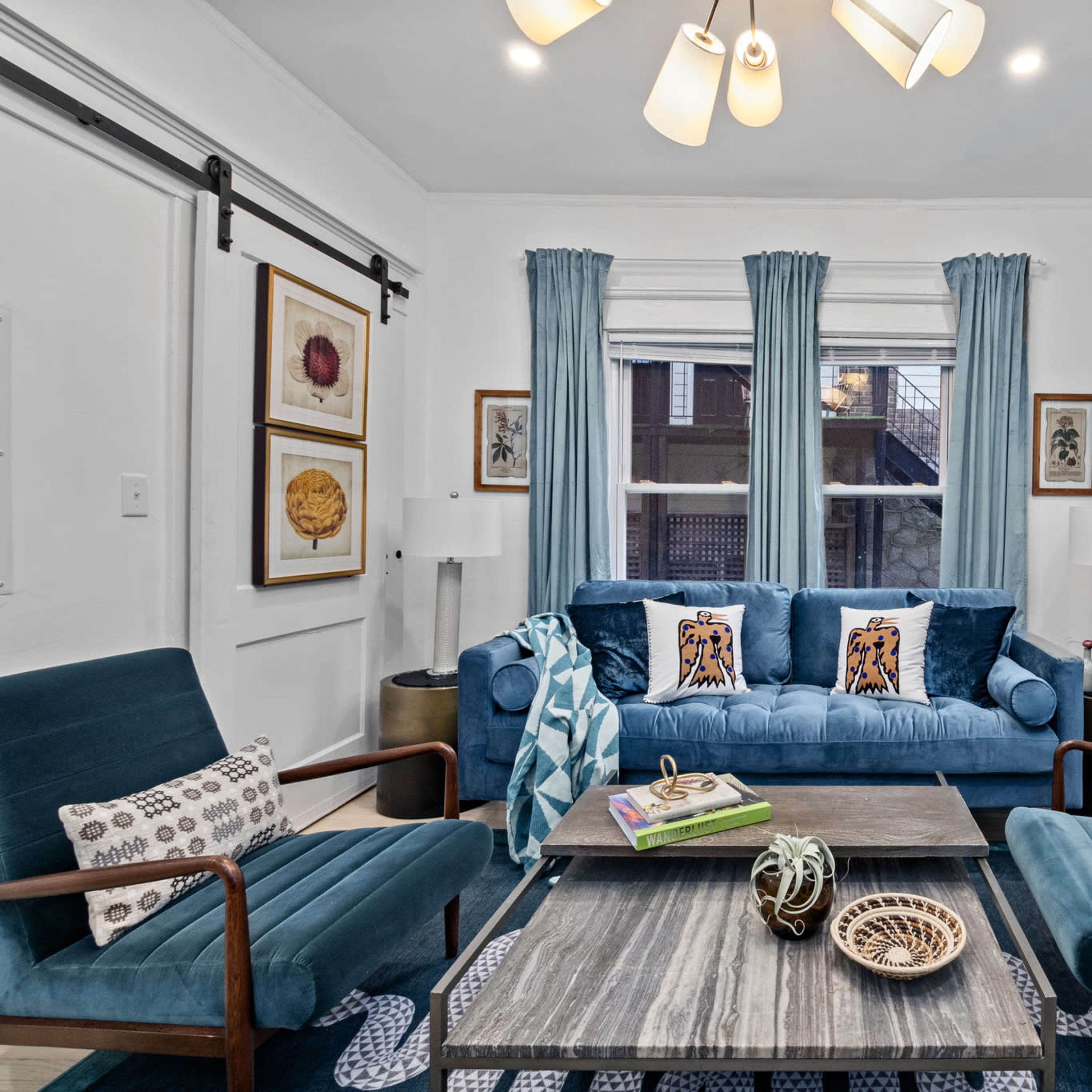A cozy living room furnished with a blue sofa, two wooden chairs, and a coffee table, with large windows dressed in light blue curtains.