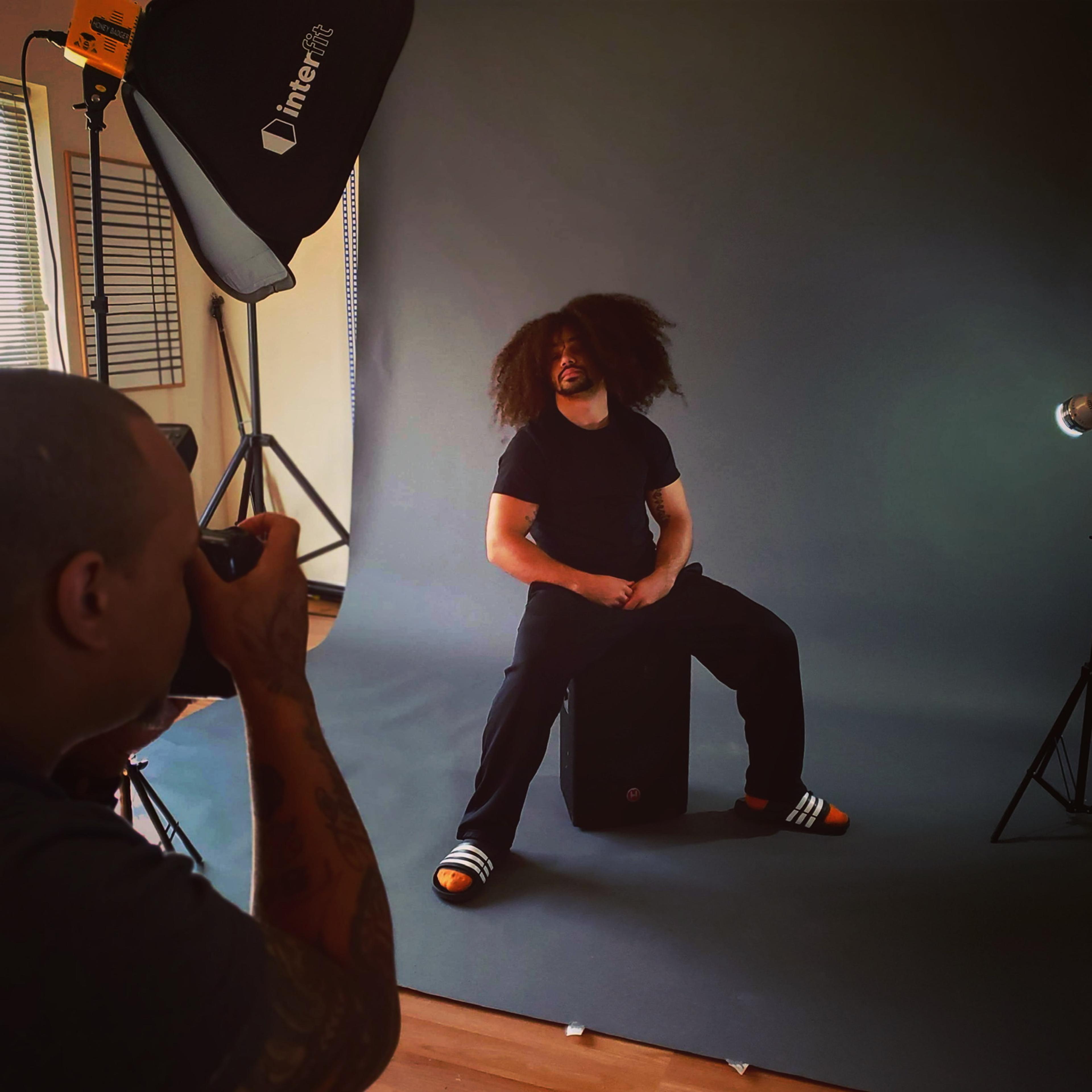A person with voluminous hair sits on a stool in front of a gray backdrop while another person photographs him.