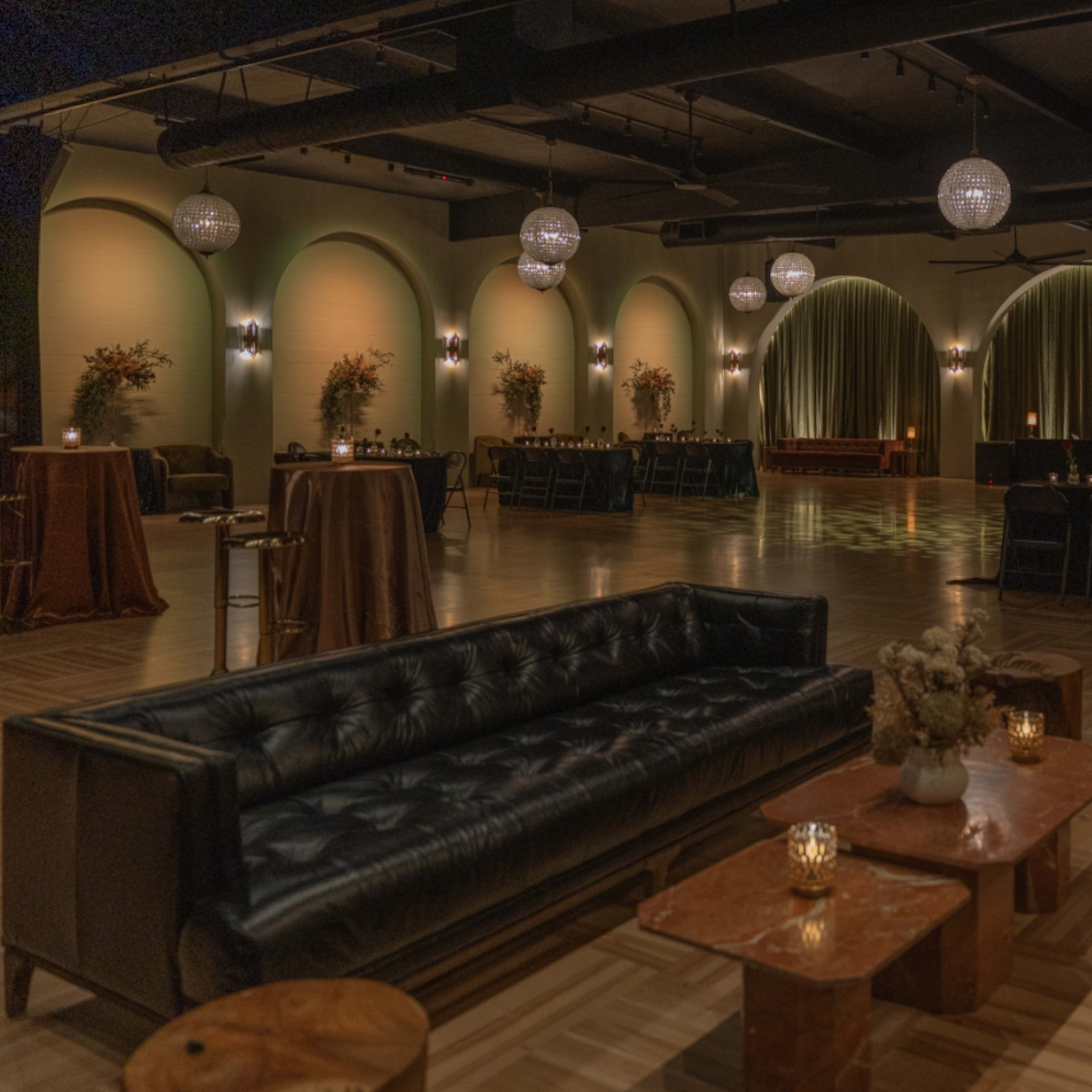An elegantly arranged event space with dark sofas, wooden tables, and ambient lighting from chandeliers and candles.