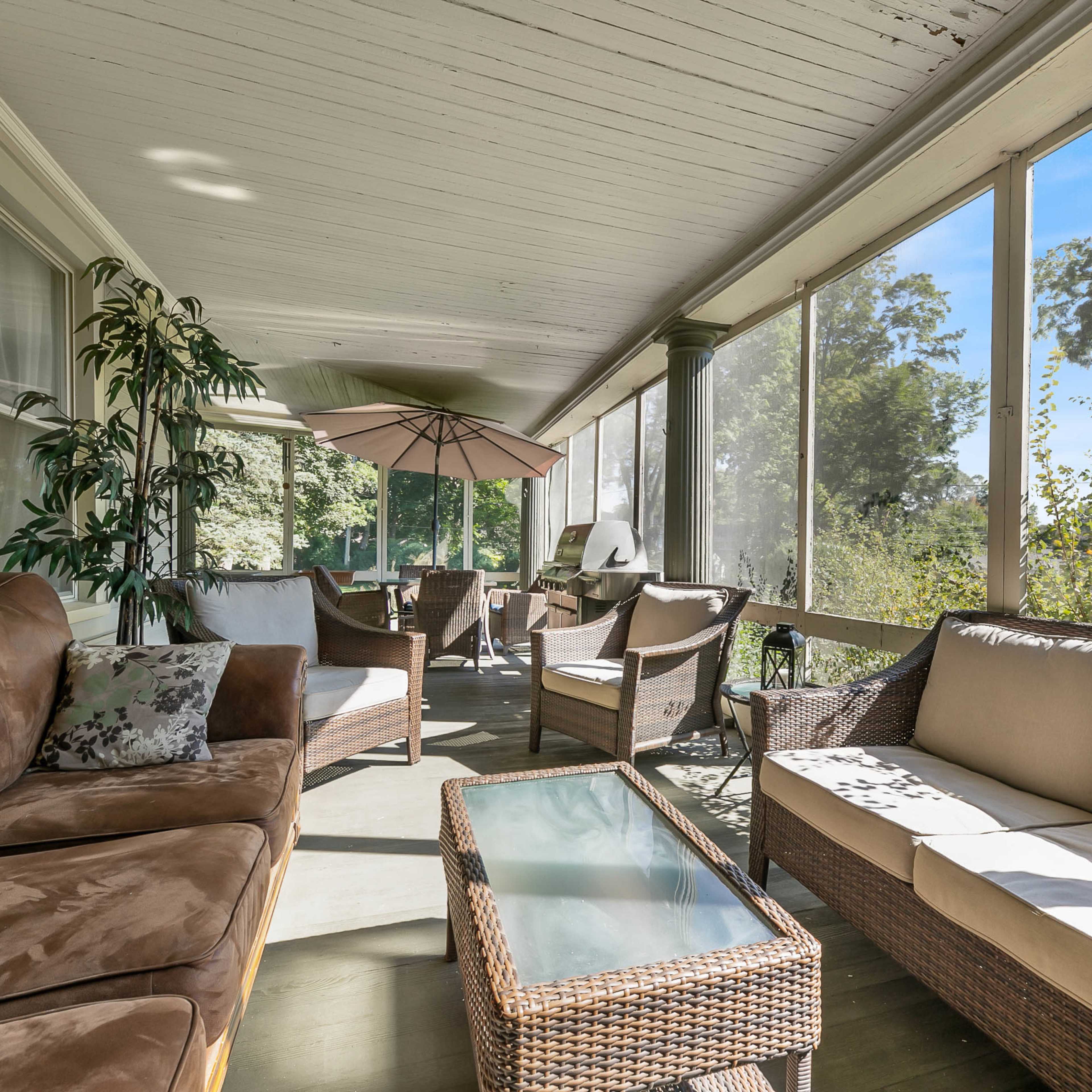 A screened porch furnished with comfortable seating, a glass coffee table, and an umbrella over a patio area overlooking a natural landscape.