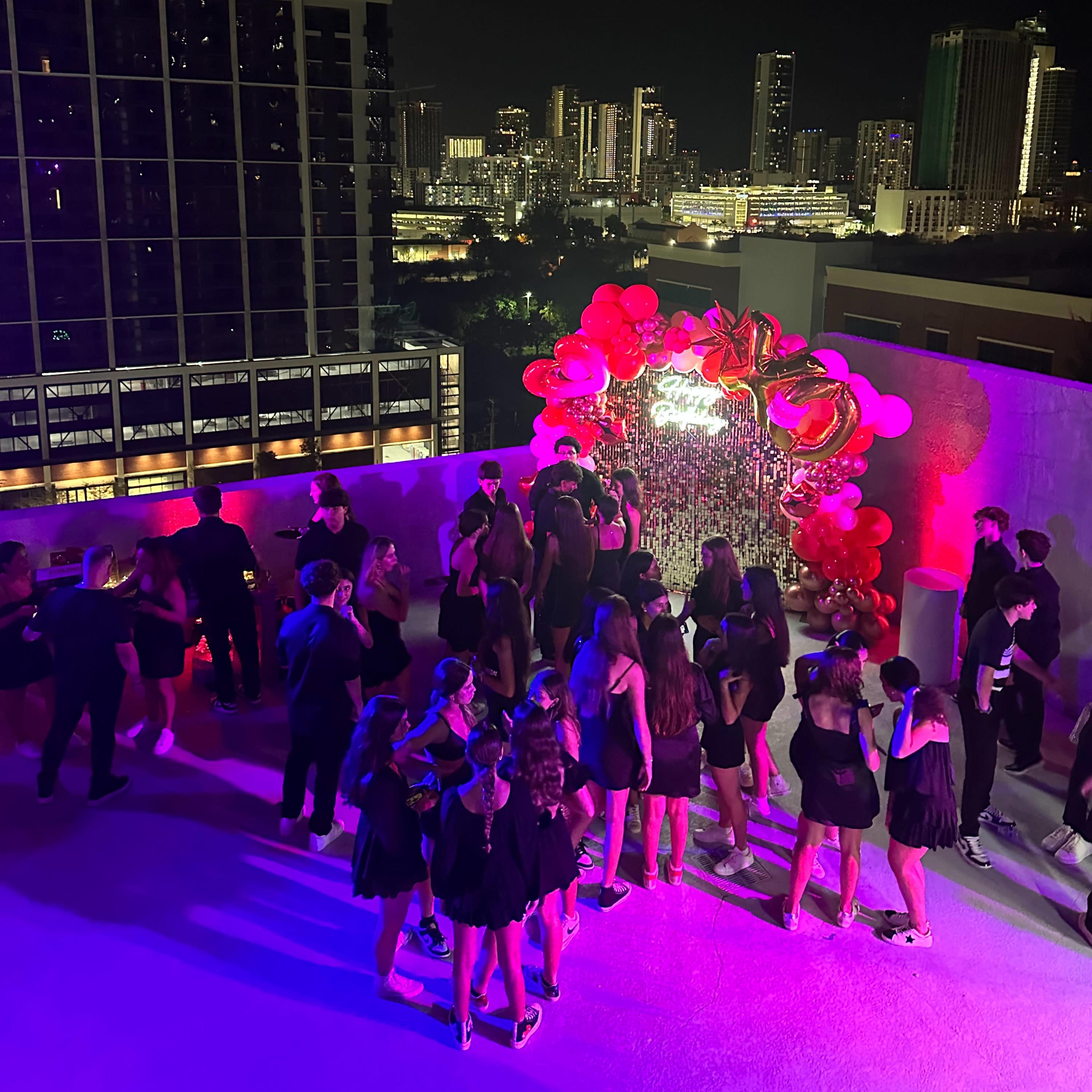 A group of people gather on a rooftop at night, surrounded by city lights and a decorative arch made of red balloons.
