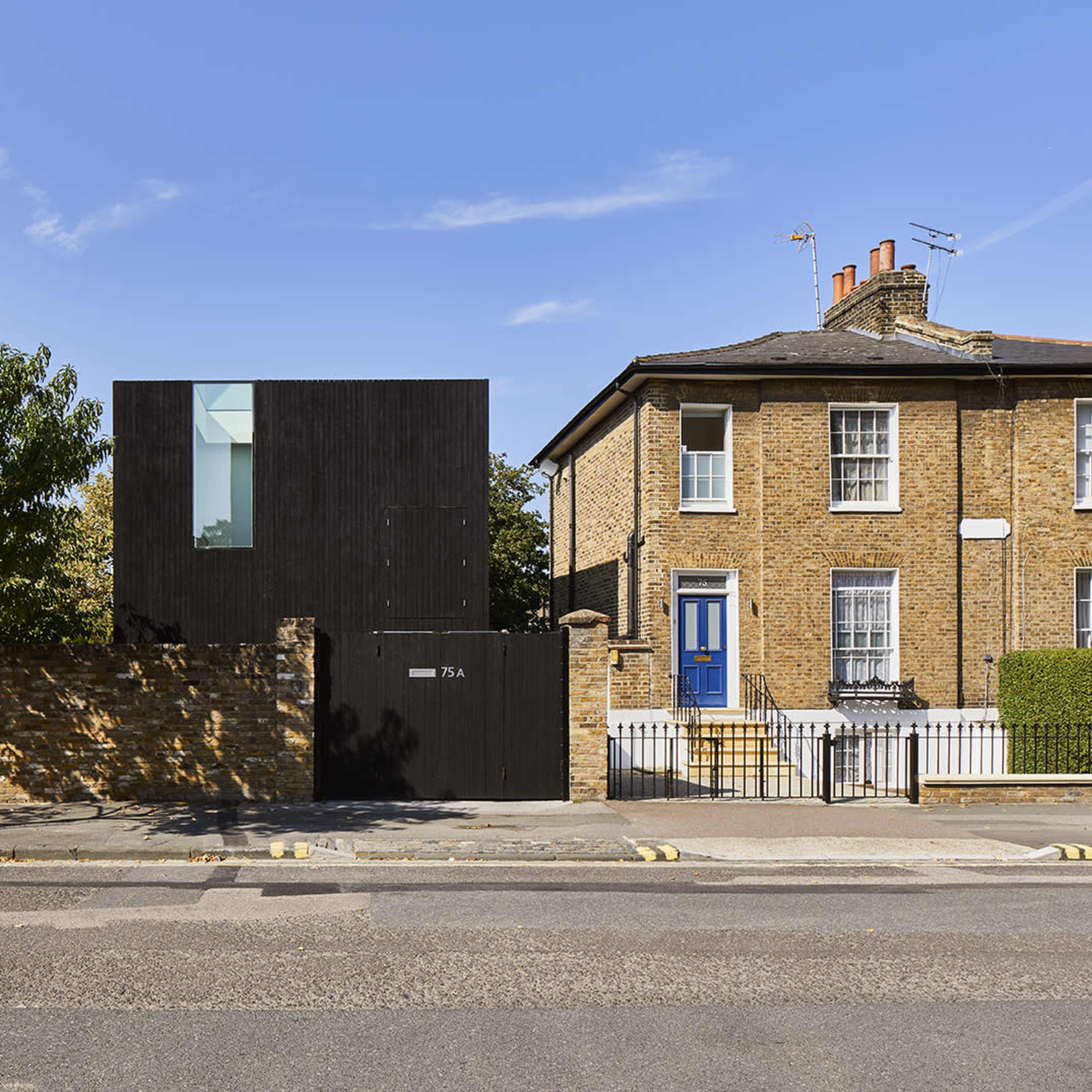 A modern black wooden house adjacent to a traditional brick house, separated by a low brick wall and street.