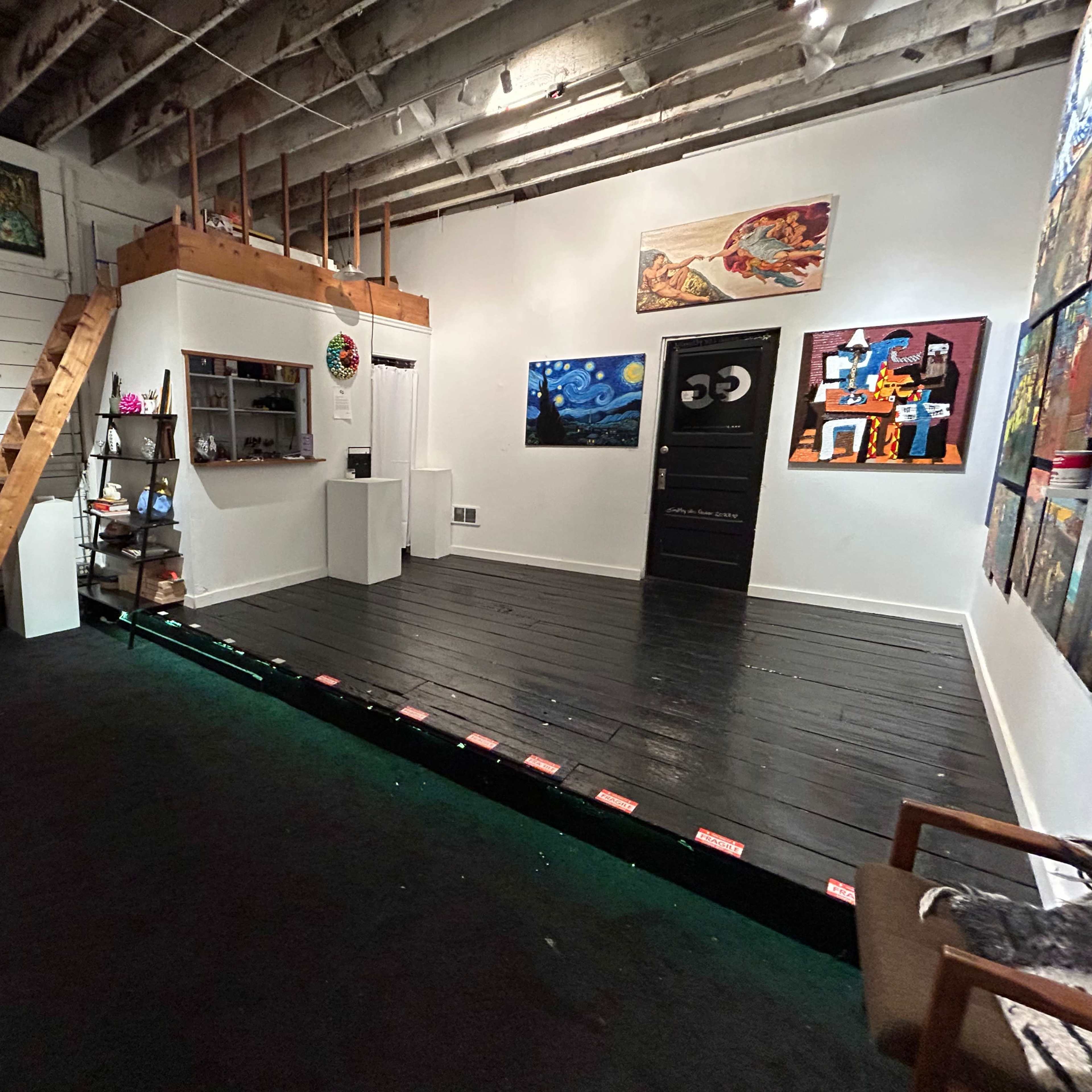A spacious art studio with a wooden staircase leading to a loft, featuring various artworks displayed on the walls and a black wooden floor.