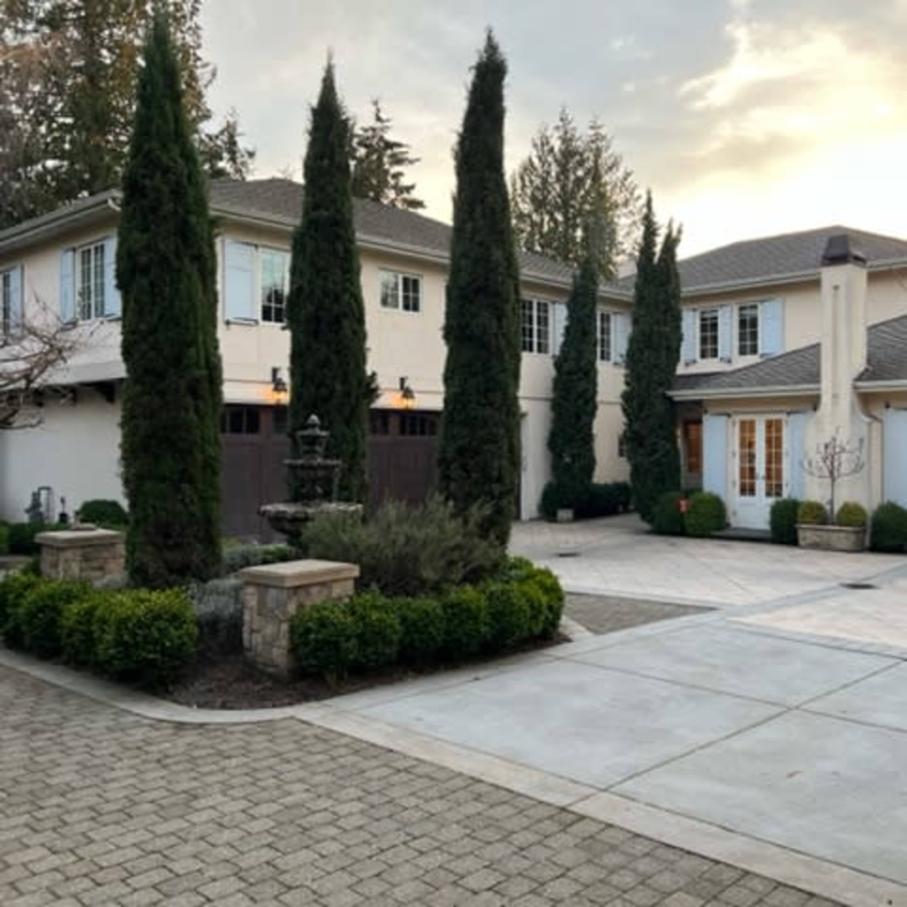 A large, two-story house with manicured landscaping, featuring tall conifer trees flanking the entrance and a stone paved driveway leading to the garage.