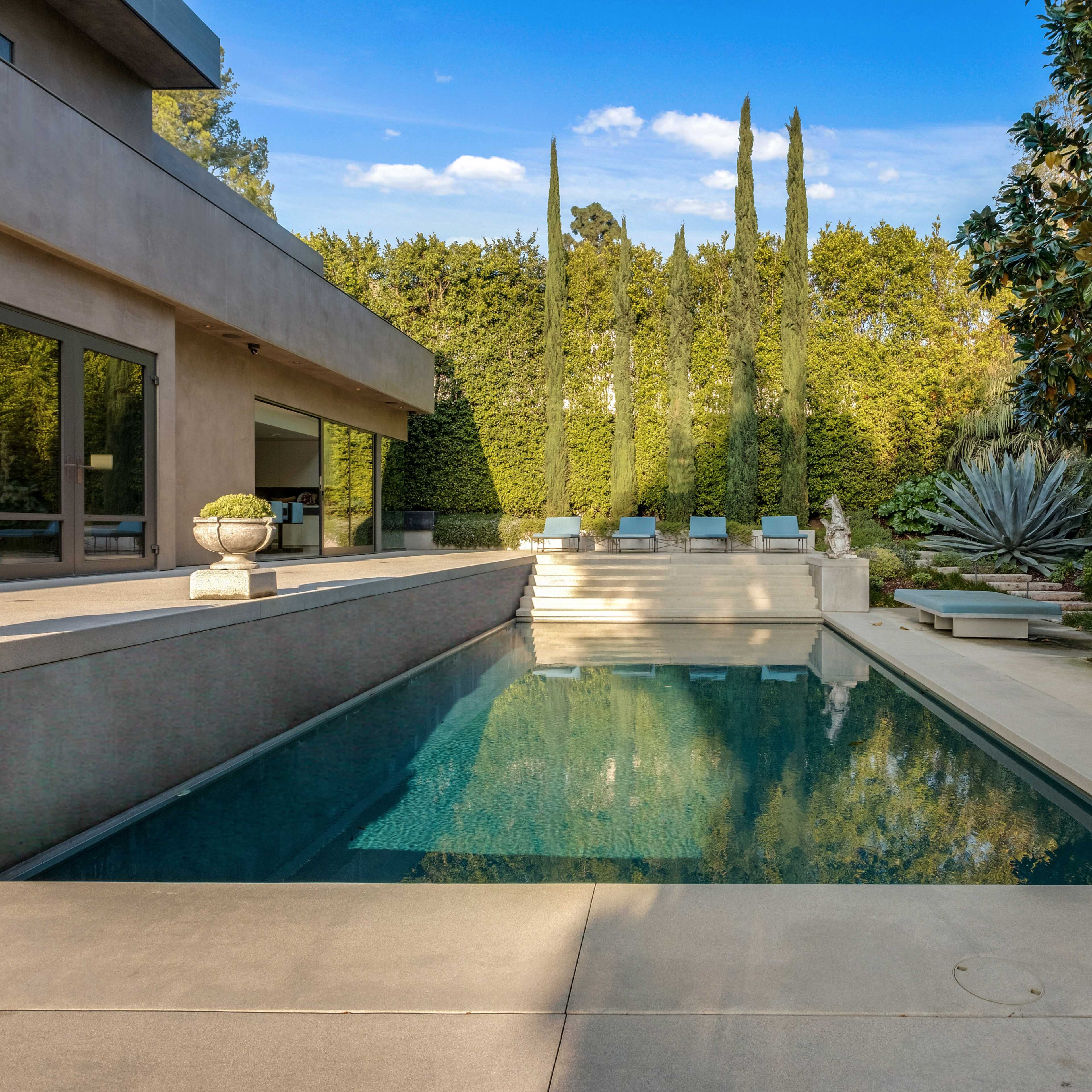 A modern pool surrounded by greenery and tall cypress trees, with a minimalistic patio area and lounge chairs.