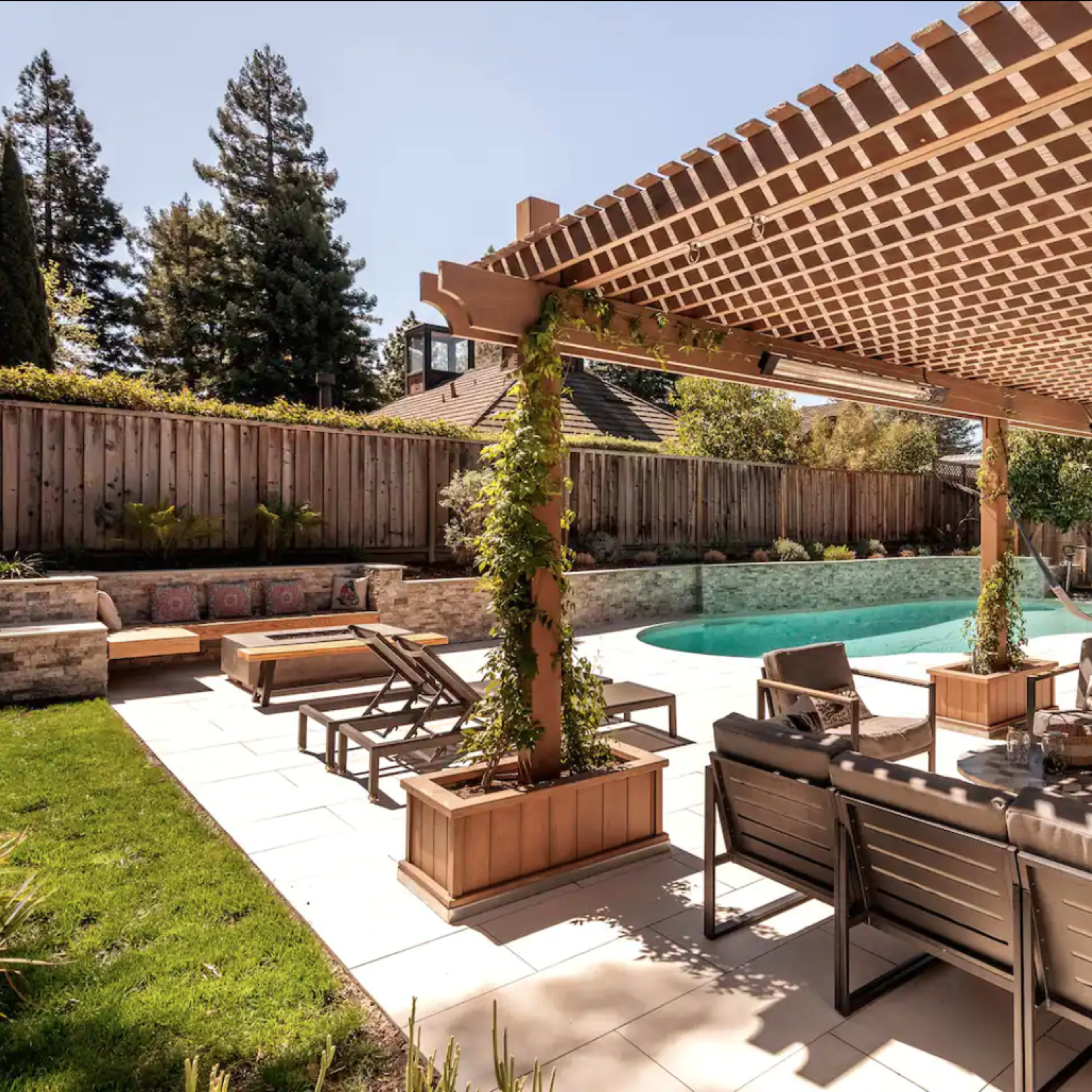 A landscaped backyard with a pool, surrounded by seating areas and a pergola covered in greenery.