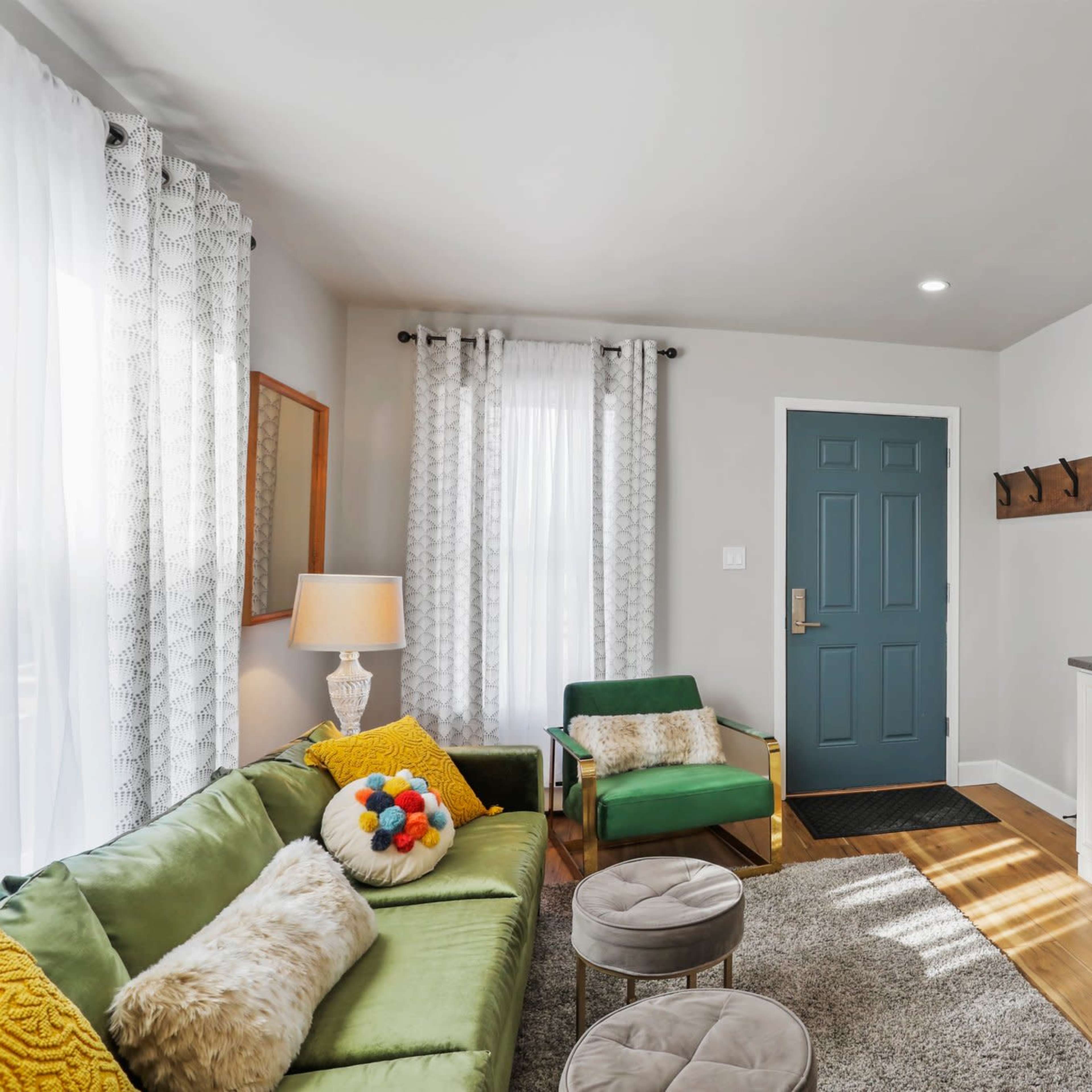 A modern living room with a green sofa, a television mounted on the wall, and a blue front door leading outside.