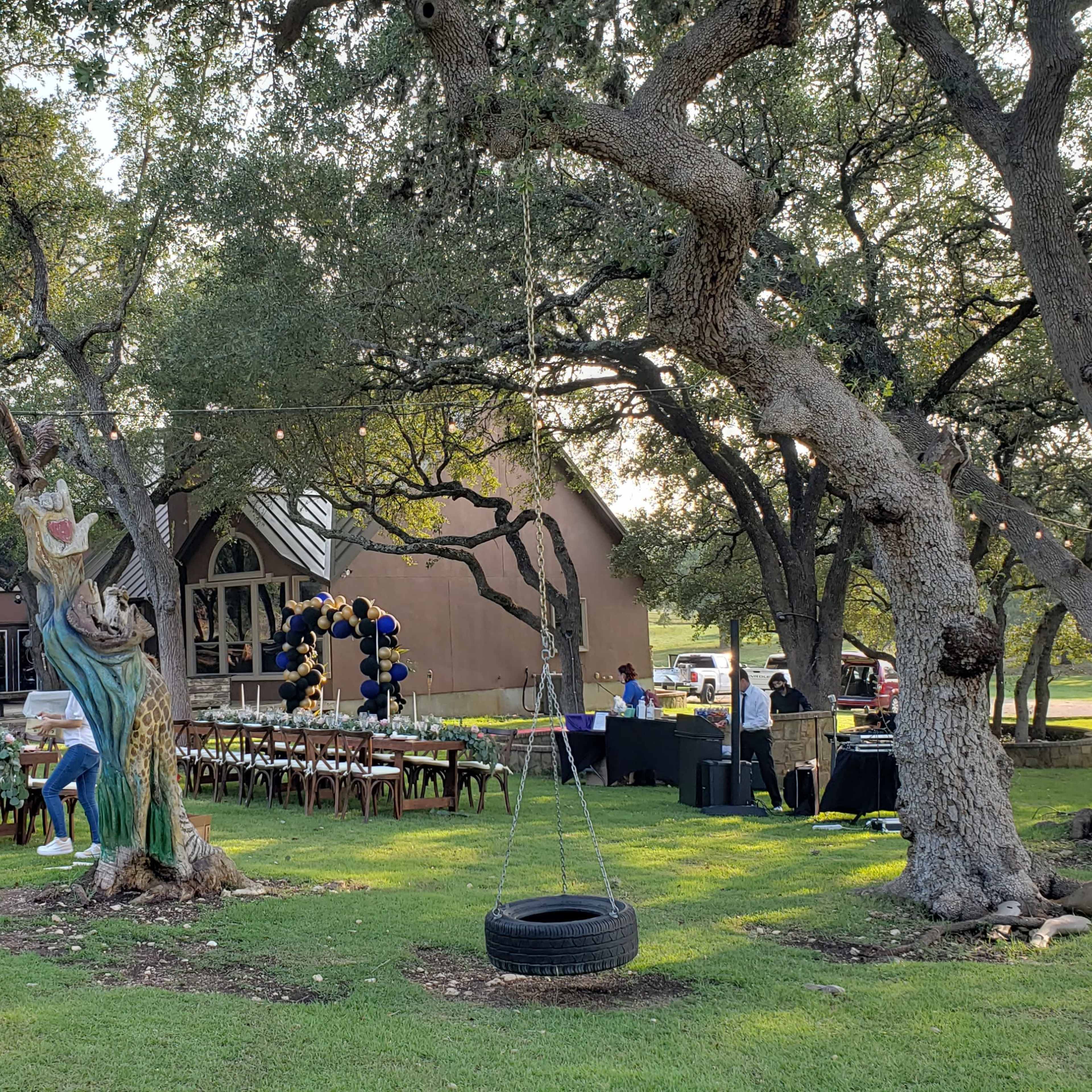 A large outdoor event setup features a long table with chairs beneath trees, adjacent to a building and decorated areas with balloons.