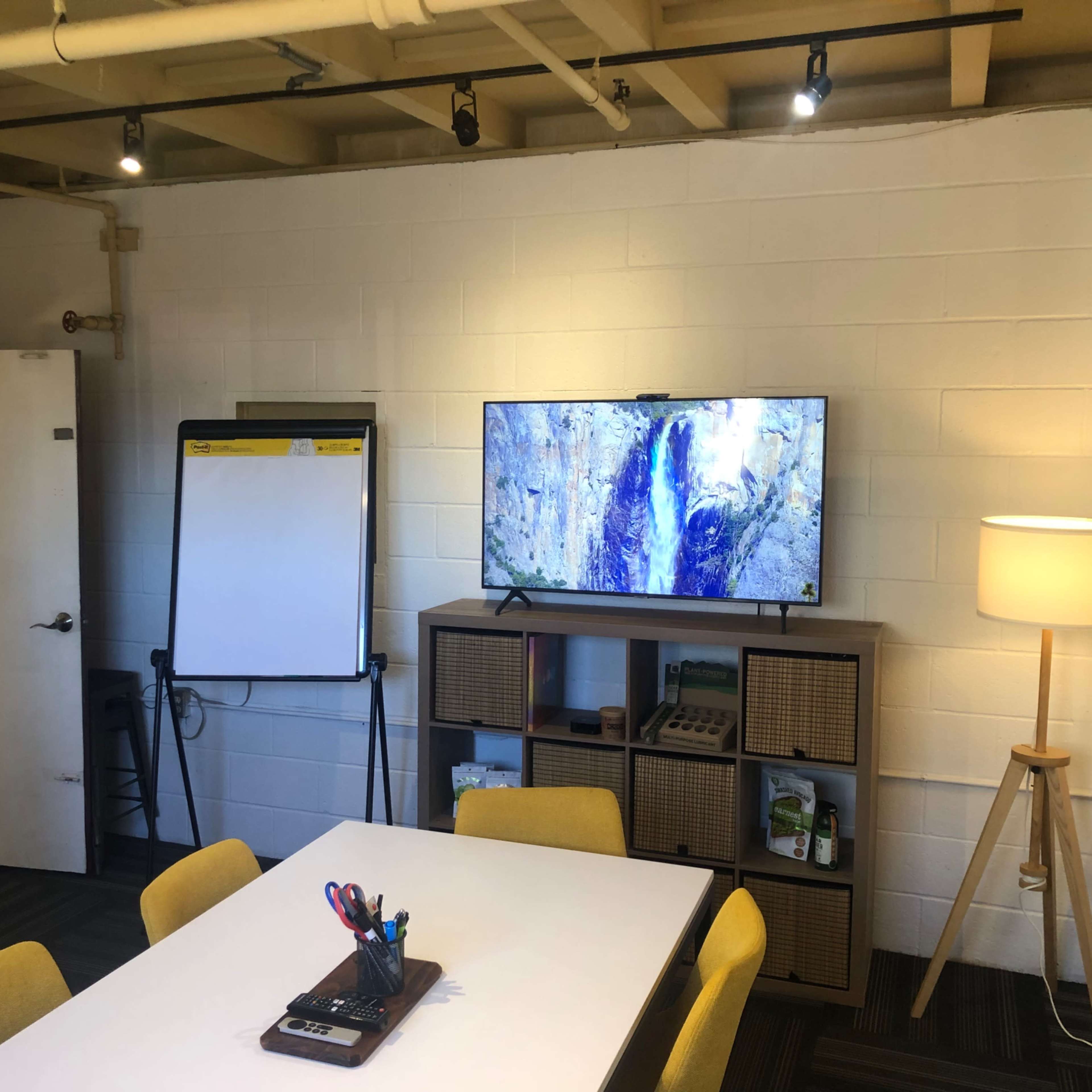 A small meeting room with a whiteboard, a television displaying a natural scene, a shelving unit, a table with pens and notebooks, and a floor lamp.
