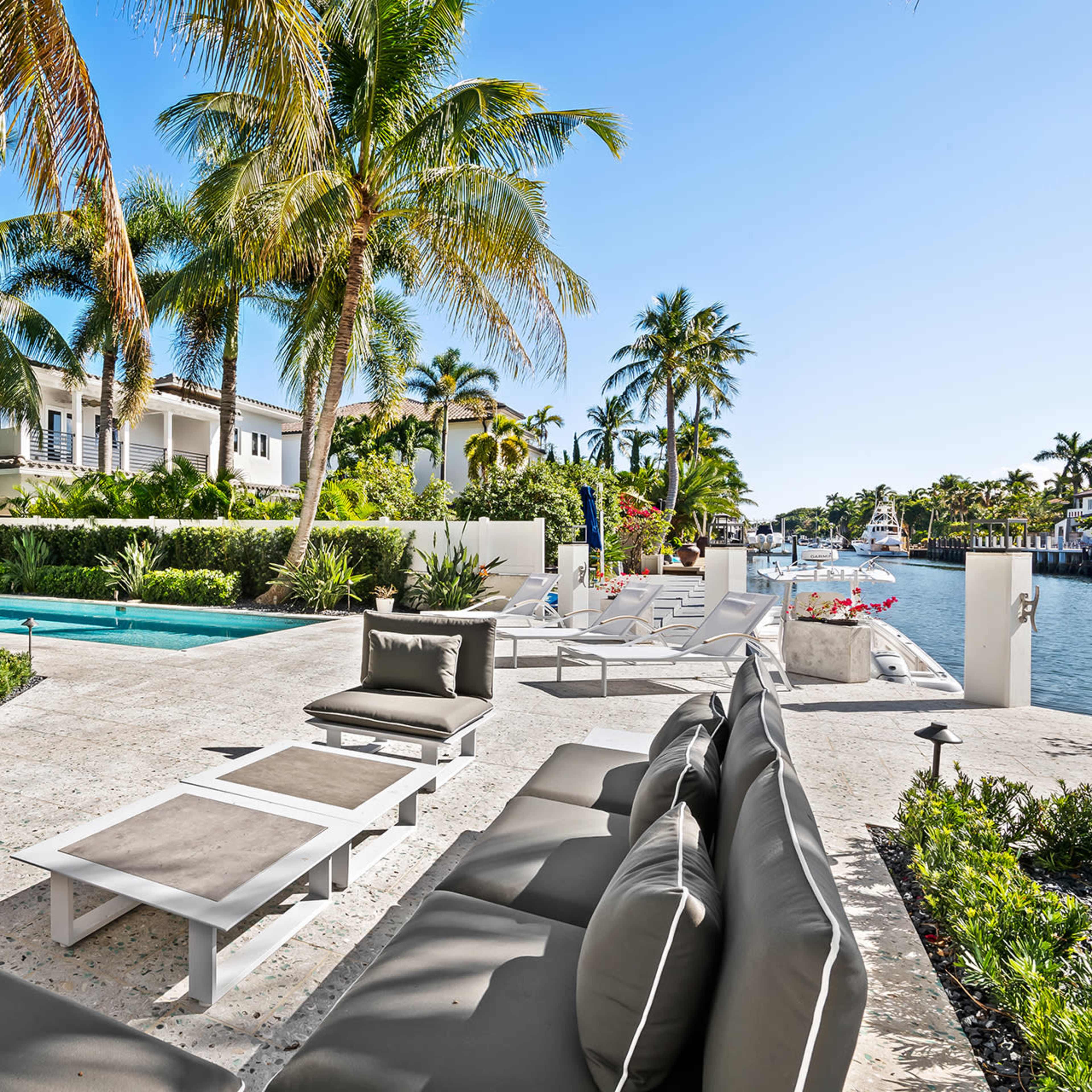 A luxurious outdoor area with a swimming pool, lounge seating, and palm trees lining a waterfront property.