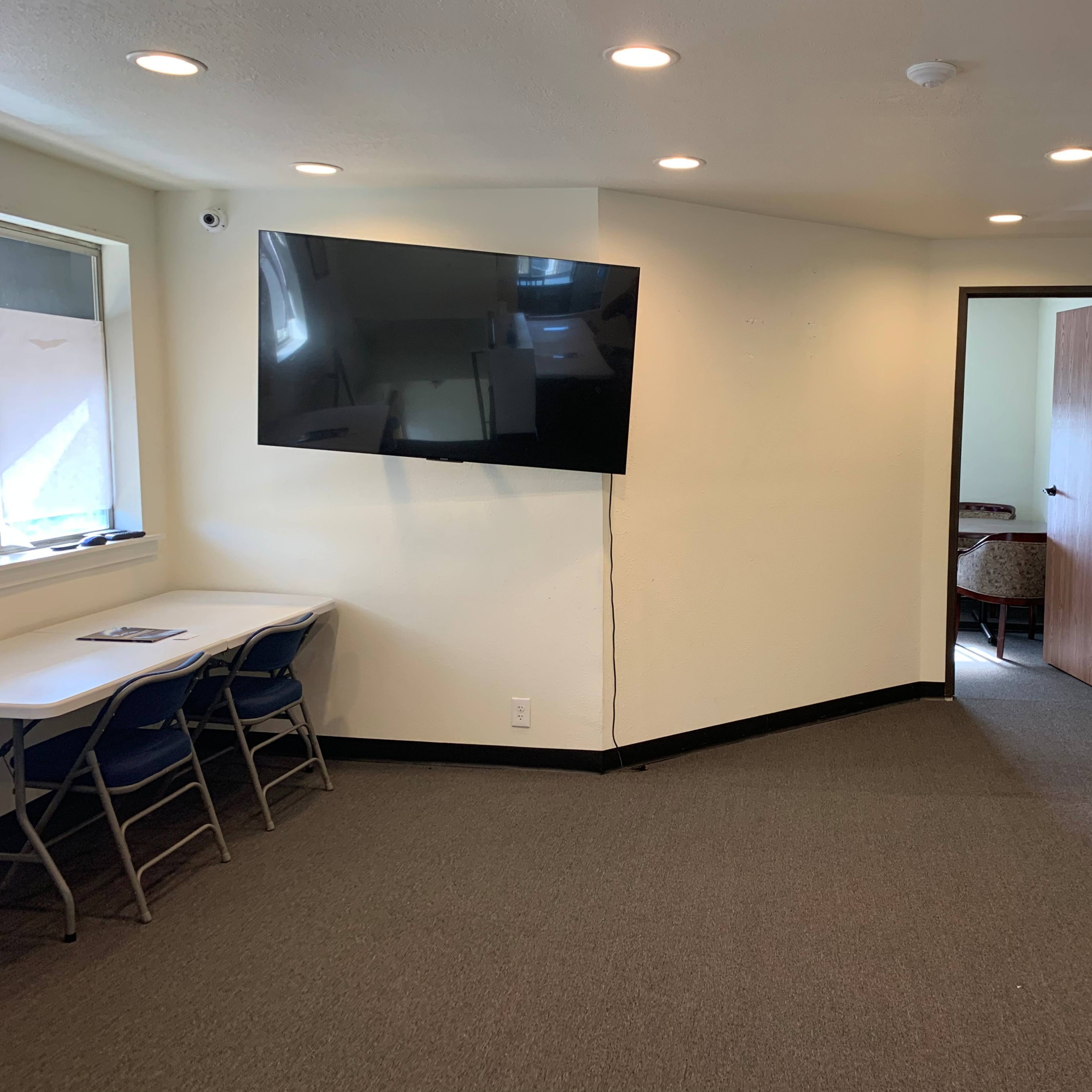 A small meeting room with a large wall-mounted TV, a table with chairs, and a door leading to another room.