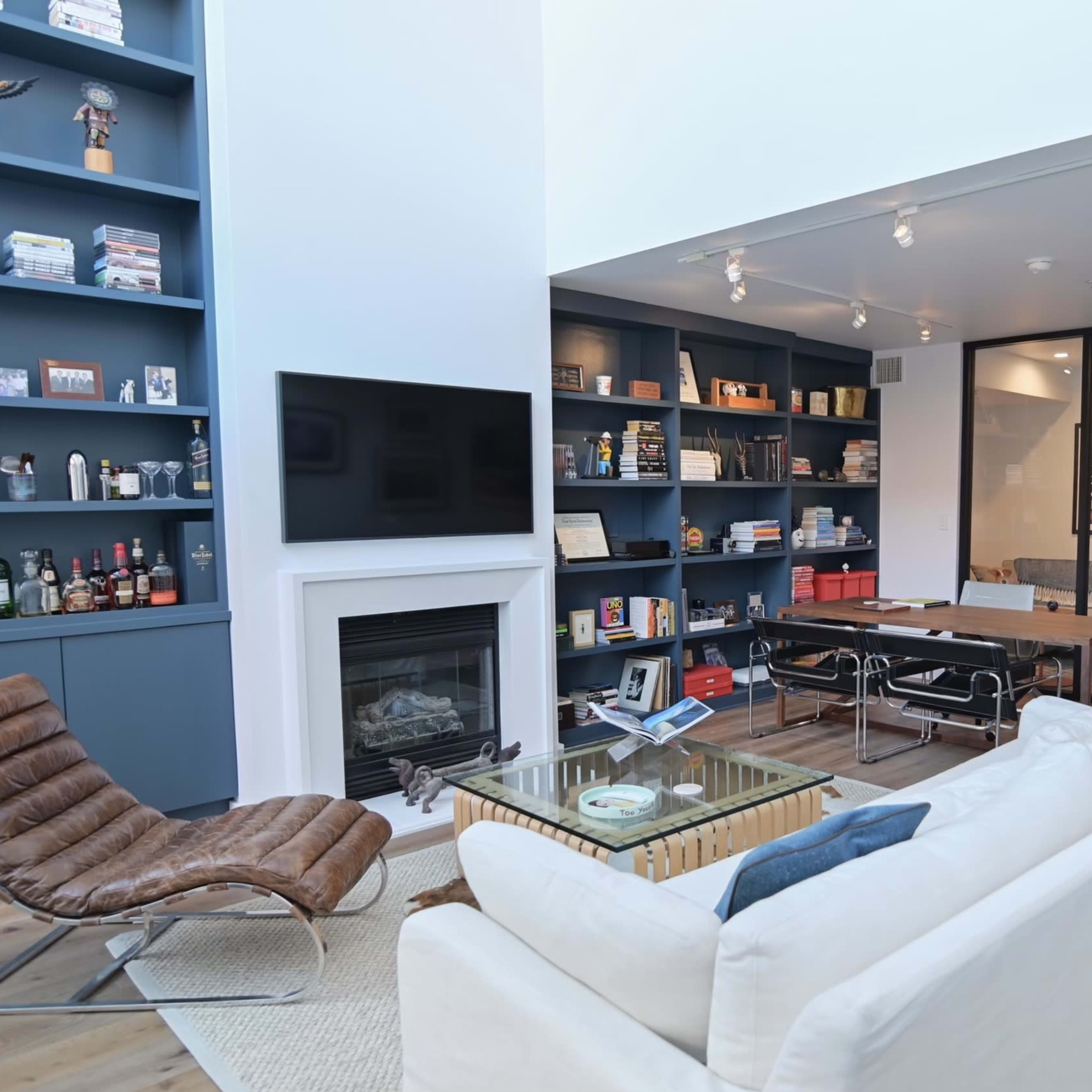 A modern living area featuring a white sofa, a glass coffee table, a fireplace, and built-in shelves filled with books and decorative items.