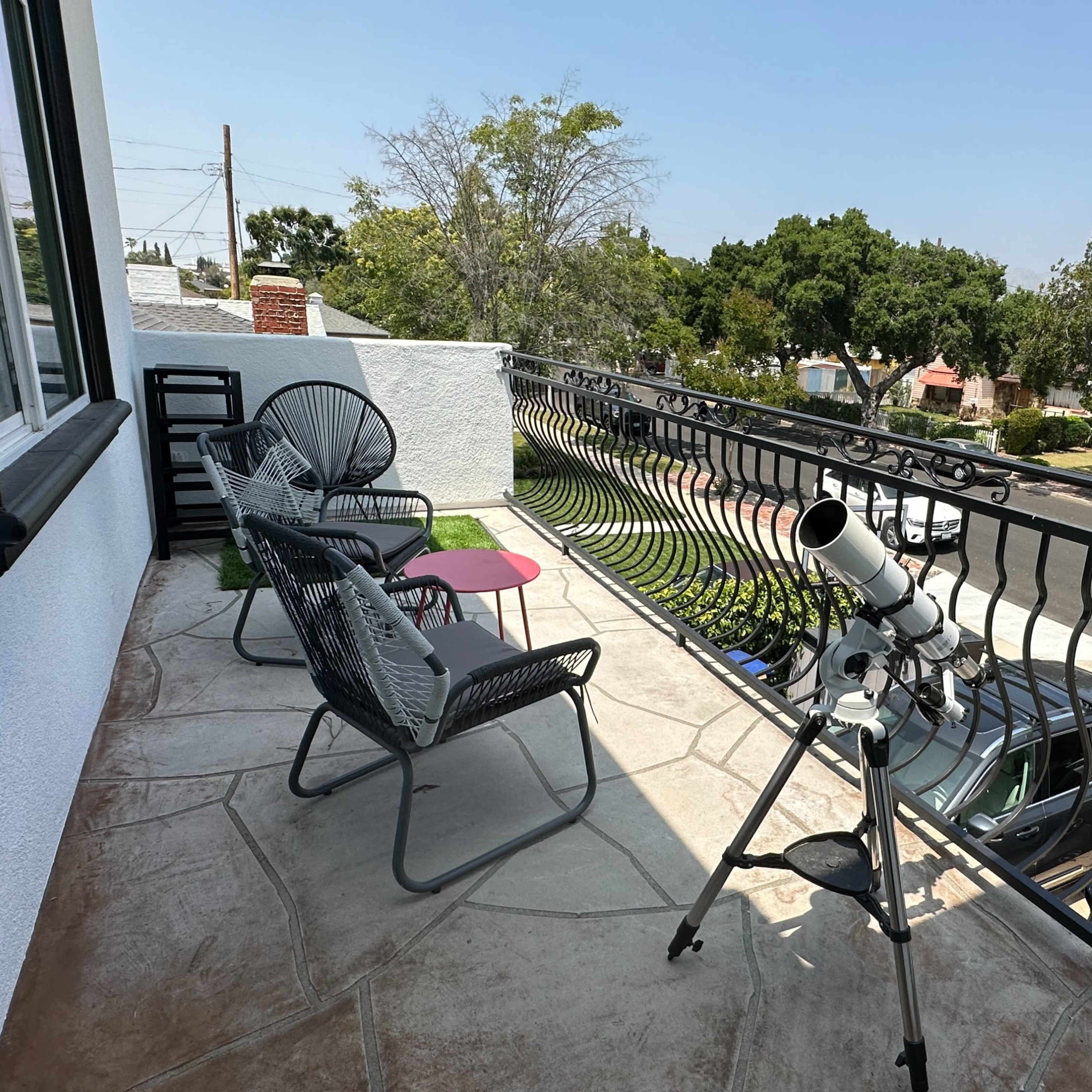 A telescope is set up on a balcony overlooking a residential street, with outdoor seating and greenery in the background.