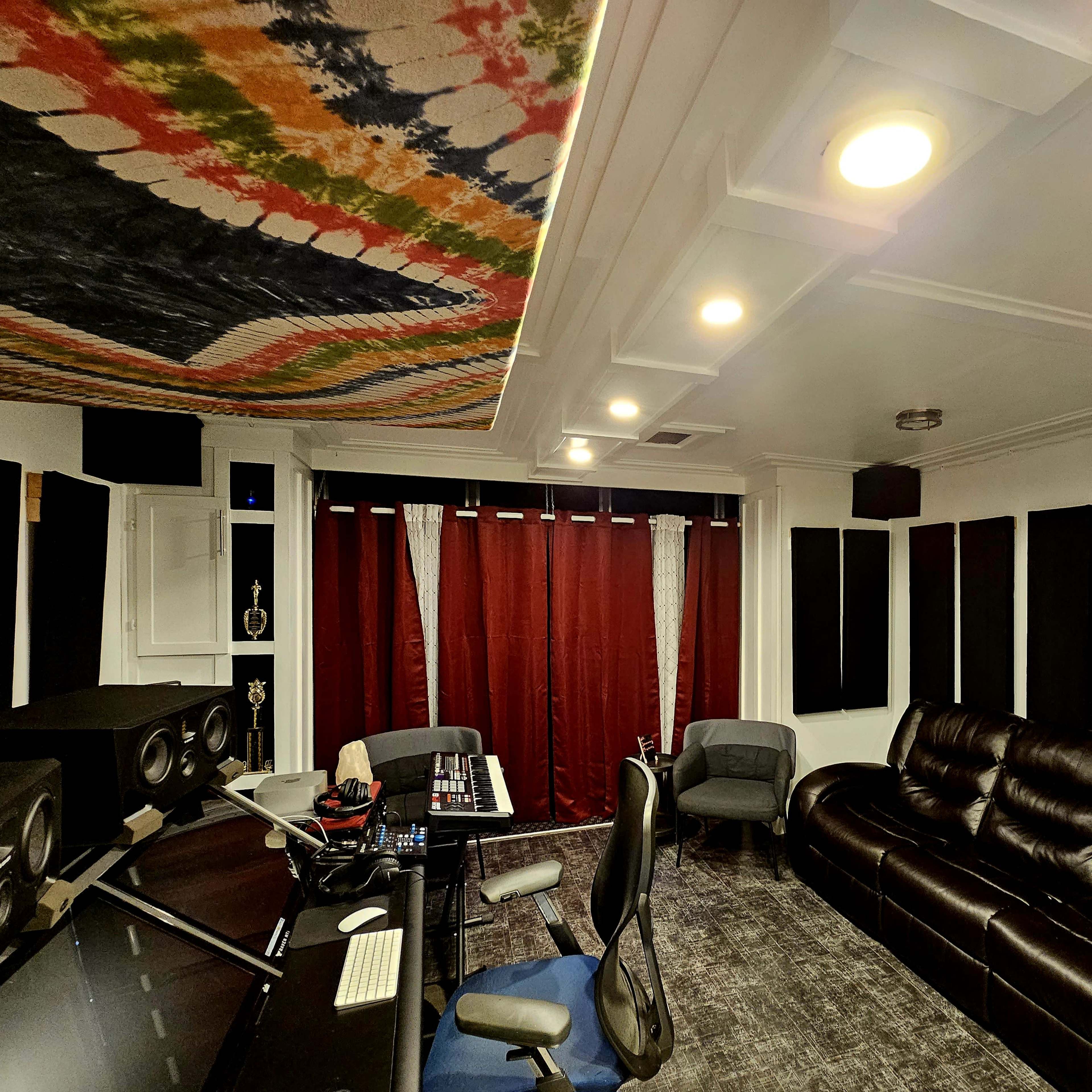 A sound studio with a black leather sofa, acoustic panels on the walls, a control desk with audio equipment, and red curtains.