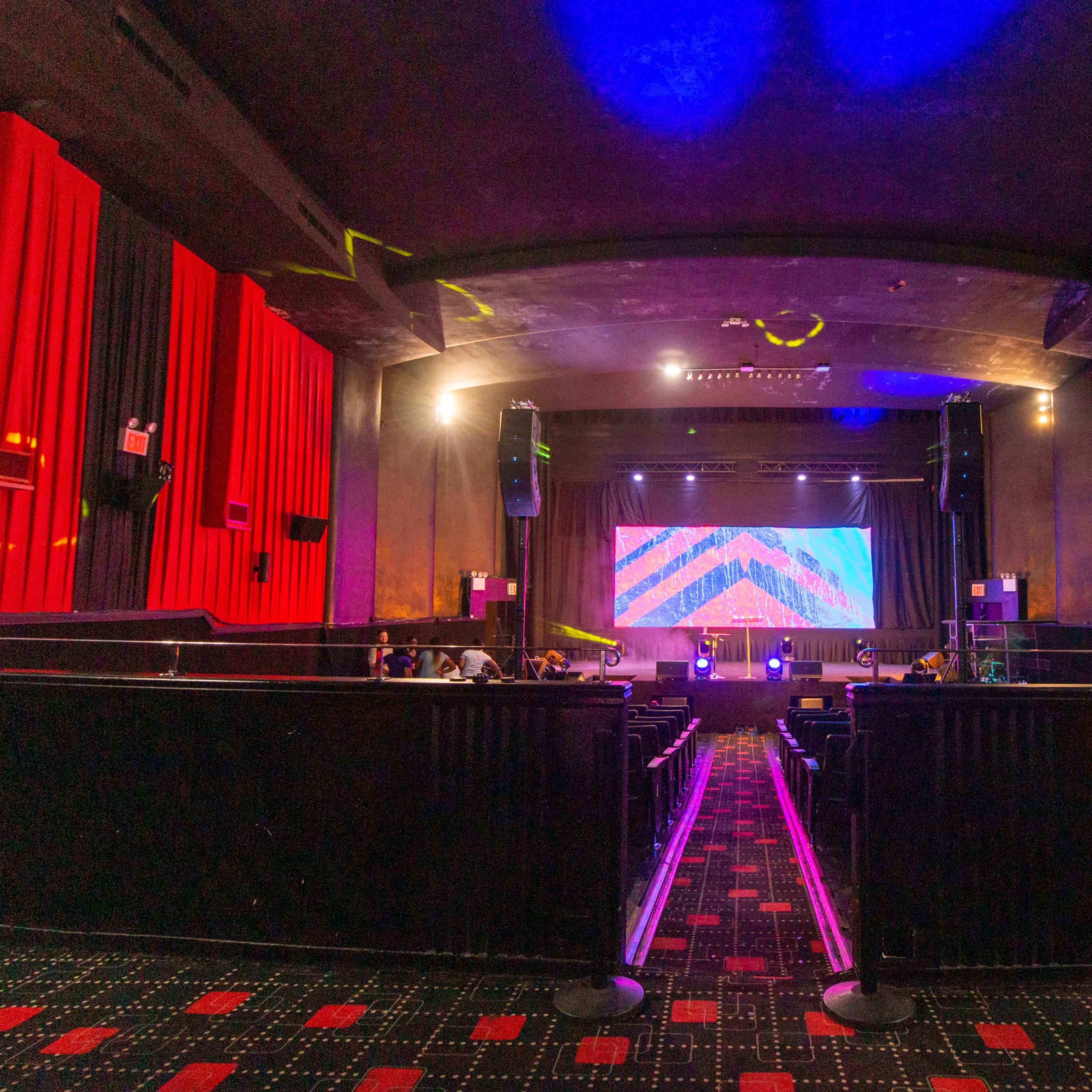 The interior of a theater with red curtains, dark seating, and a colorful stage backdrop illuminated by stage lights.