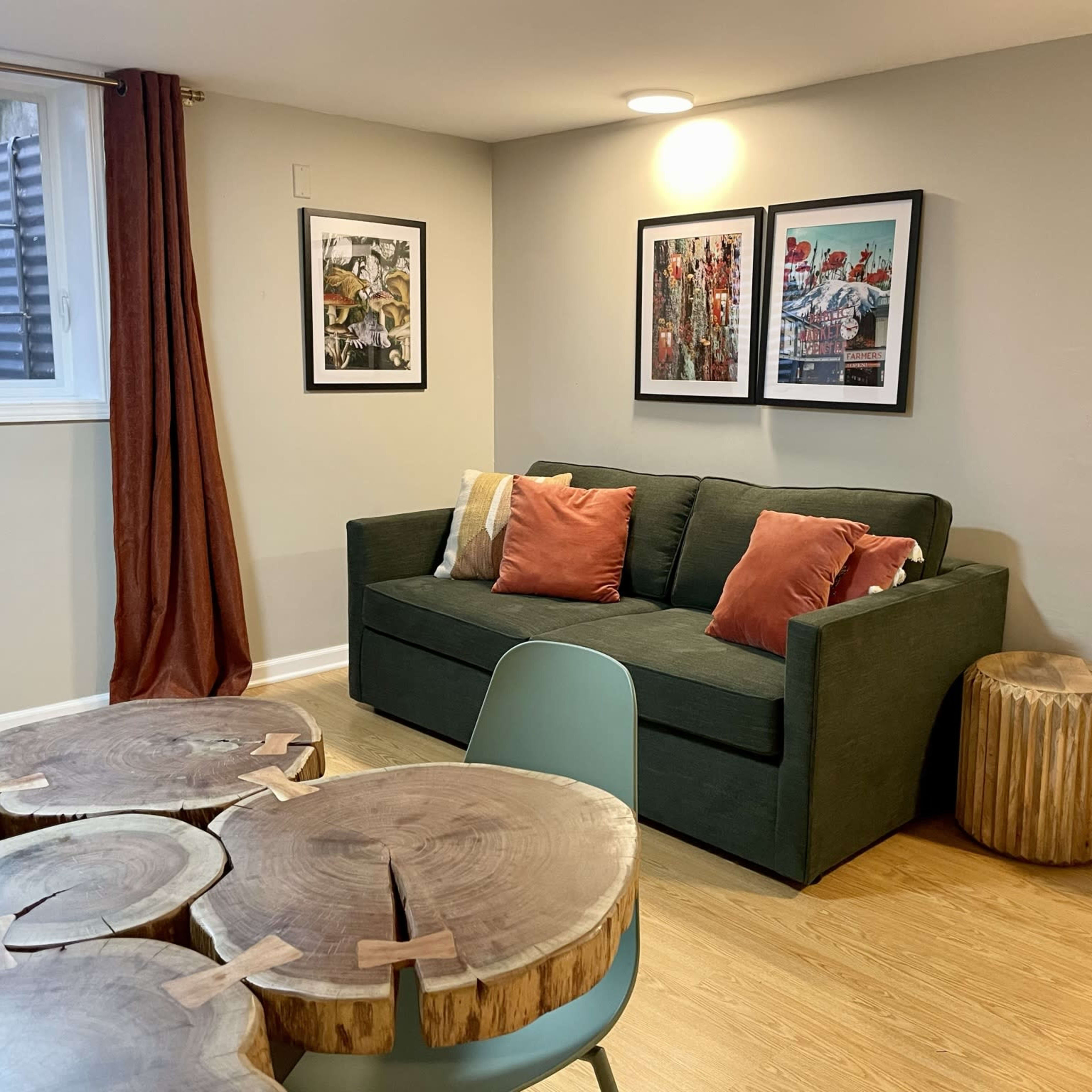 A minimalist living space with a green sofa, decorative cushions, two framed artworks on the wall, a wooden side table, and a small white microwave beside it.