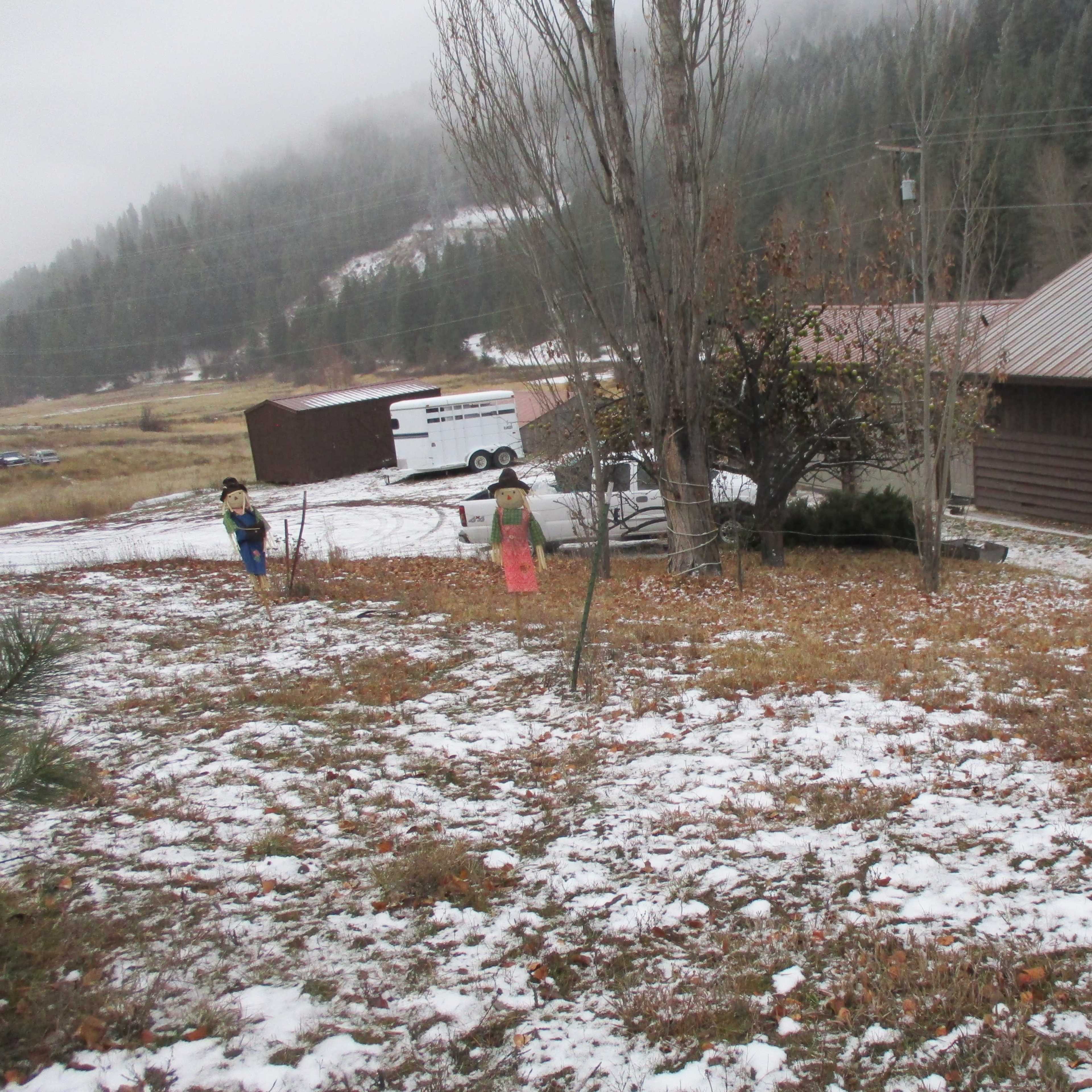 Three scarecrows stand in a yard covered with fallen leaves and patches of snow, set against a background of mountains and a wooden house.