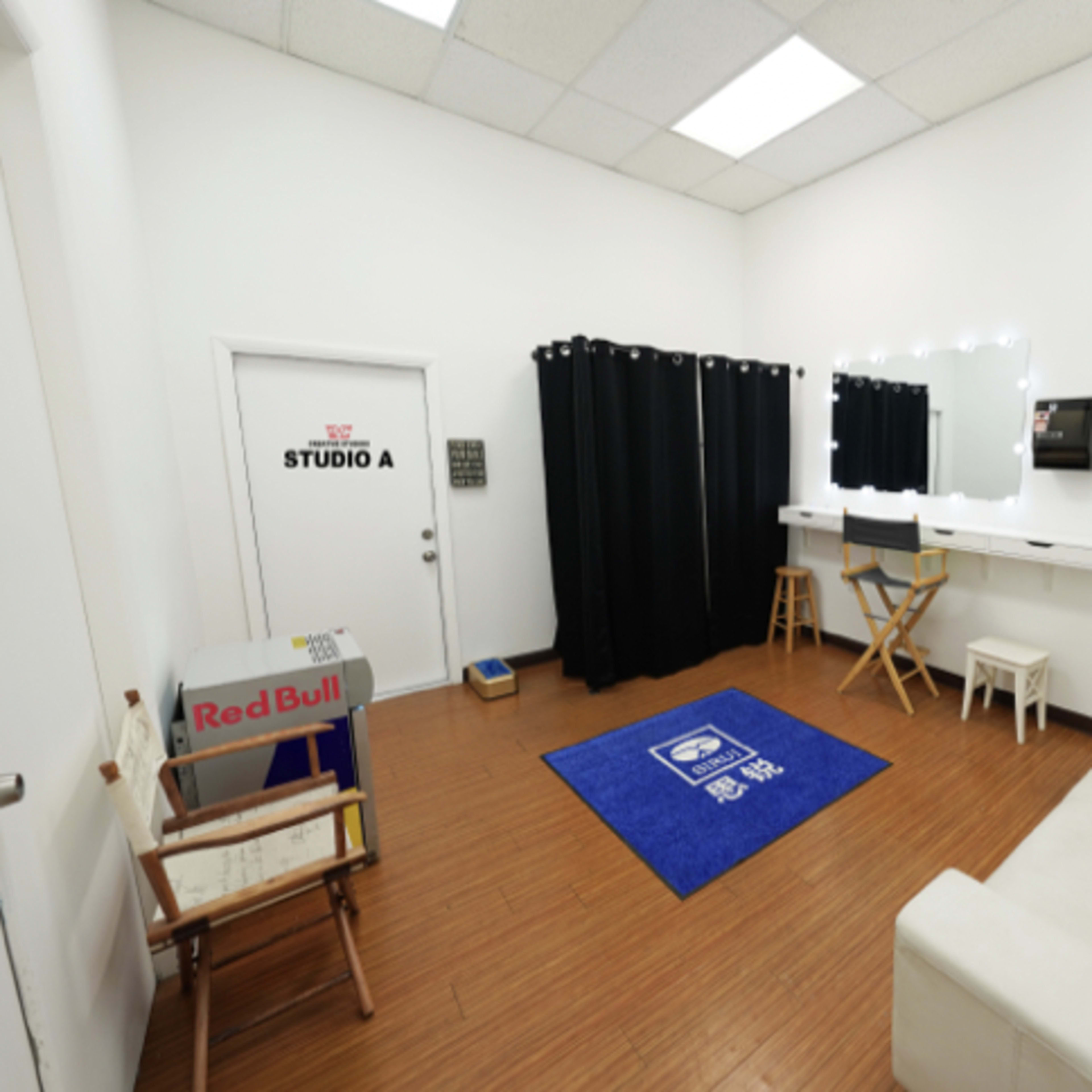 A small, modern studio room with a white sofa, a blue rug, a mirror with lights, and a curtain, along with a door labeled "STUDIO A."