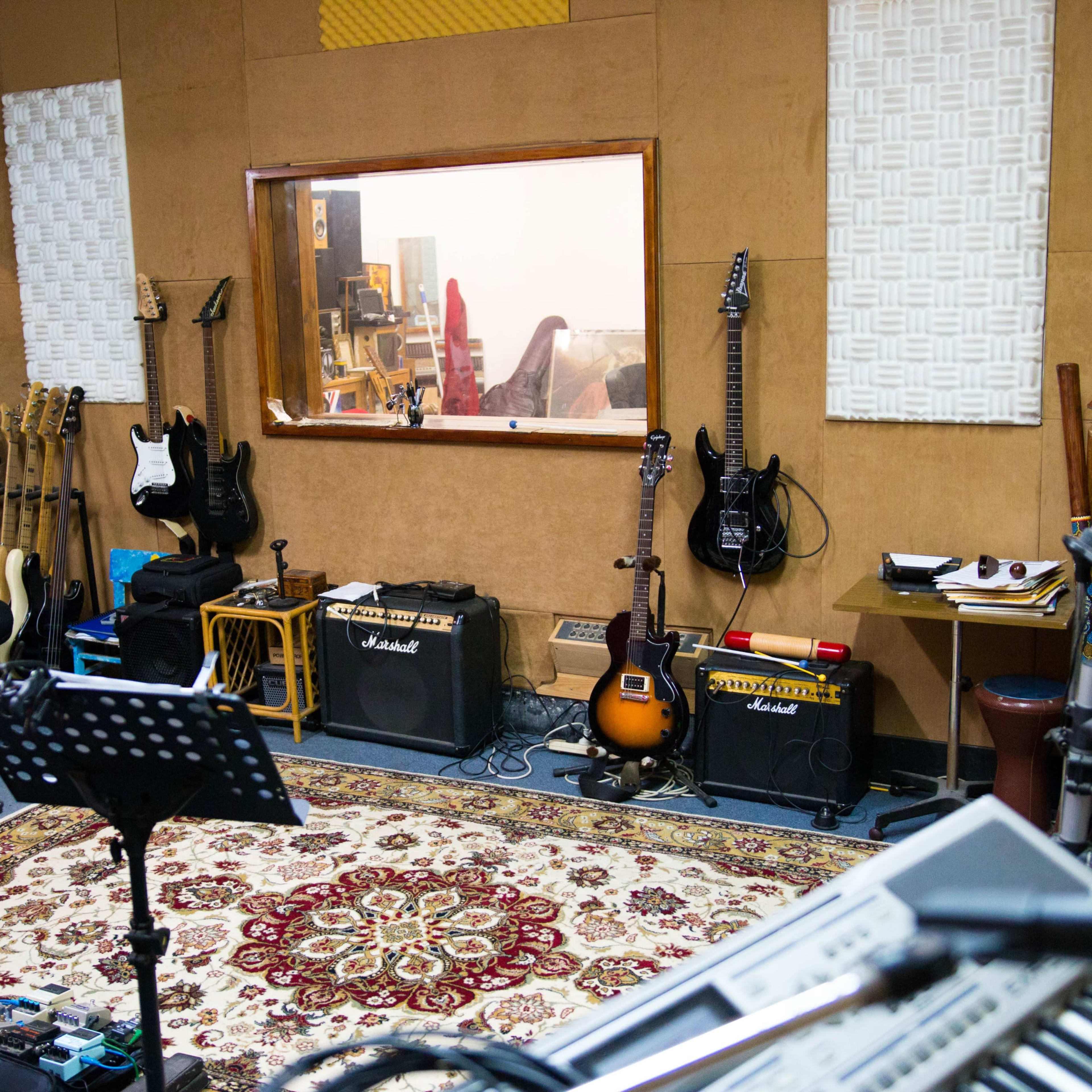 A music recording studio with various guitars, amplifiers, and a patterned rug, along with a control room visible through a window.