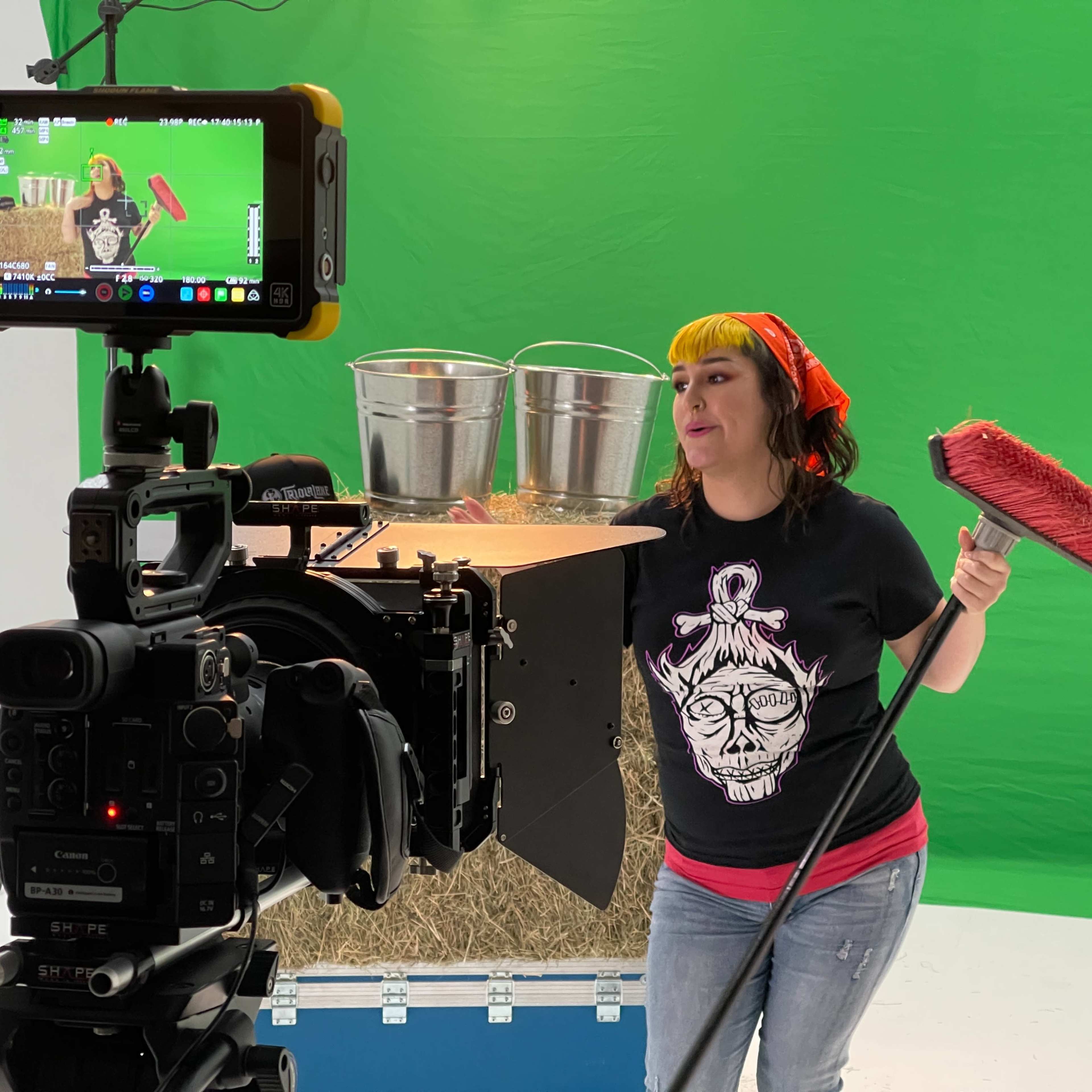 A woman in a black shirt and red bandana stands in front of a green screen, holding a broom and posing near buckets on a set with hay.