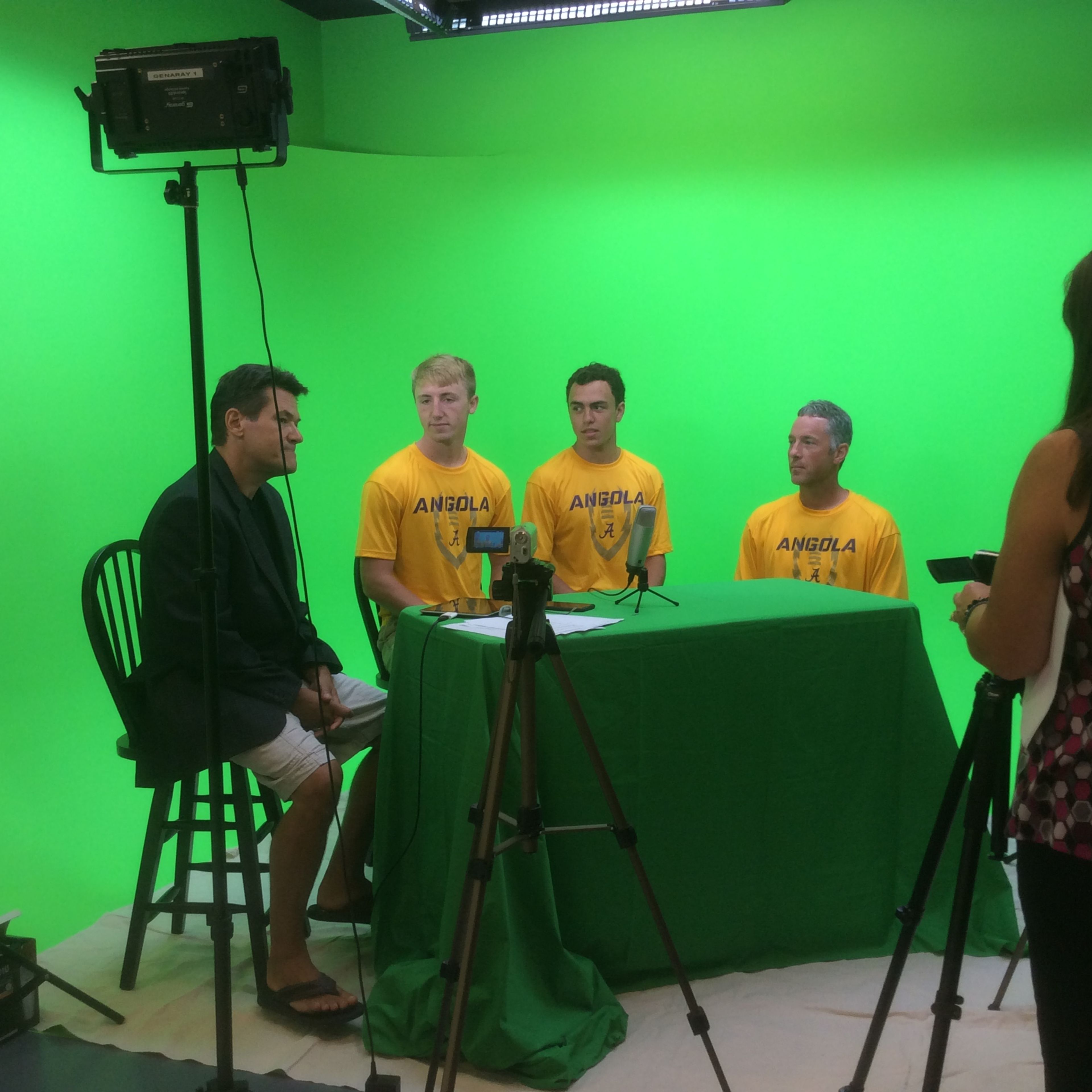 A group of four individuals, wearing yellow shirts marked "ANGOLA," is seated in front of a green screen, while a person on the right captures the scene with a camera.