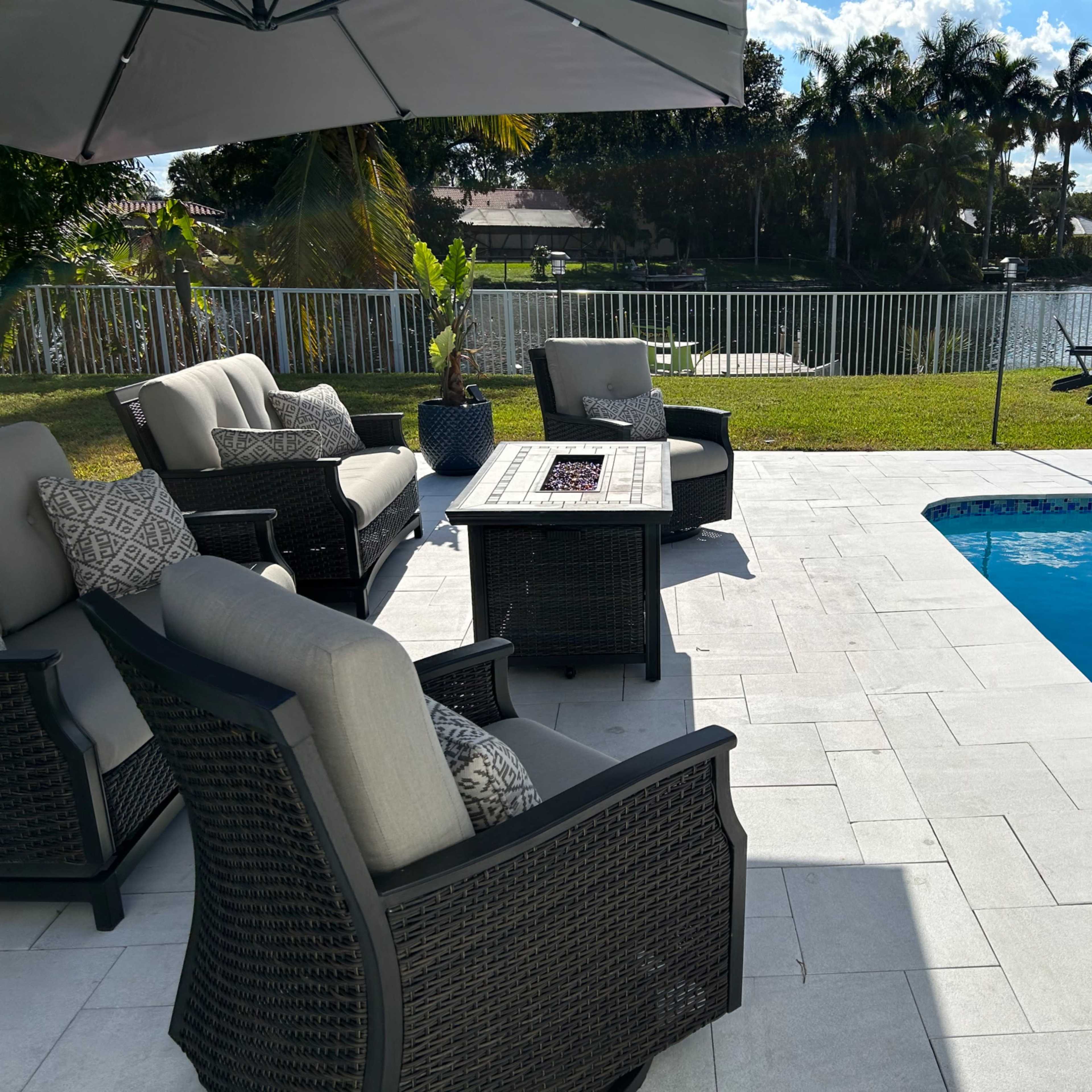 A patio area with outdoor seating, a coffee table, and a swimming pool, overlooking a grassy yard and a lake surrounded by trees.
