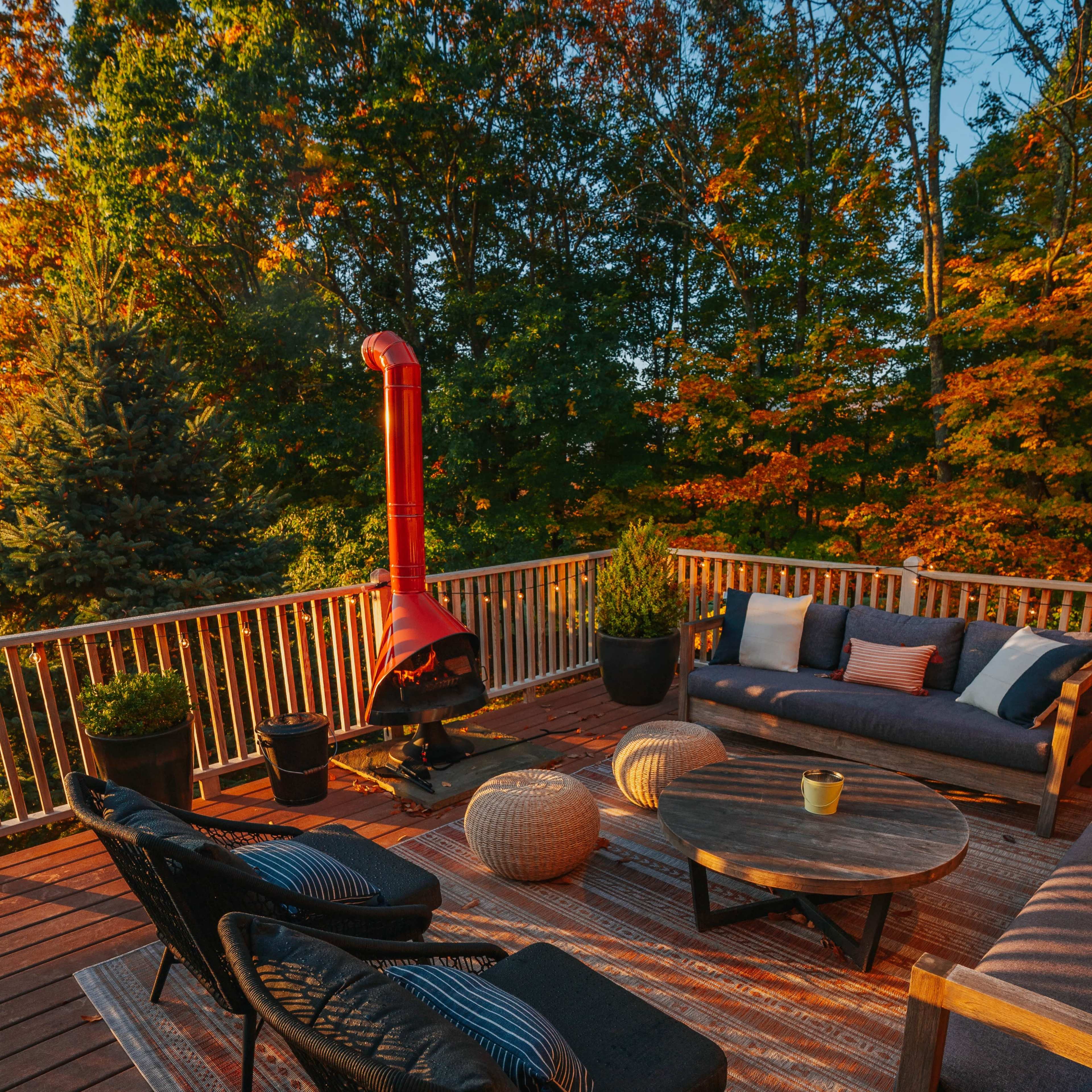 A wooden deck is furnished with modern seating, a round table, an outdoor fireplace, and surrounded by trees with autumn foliage.