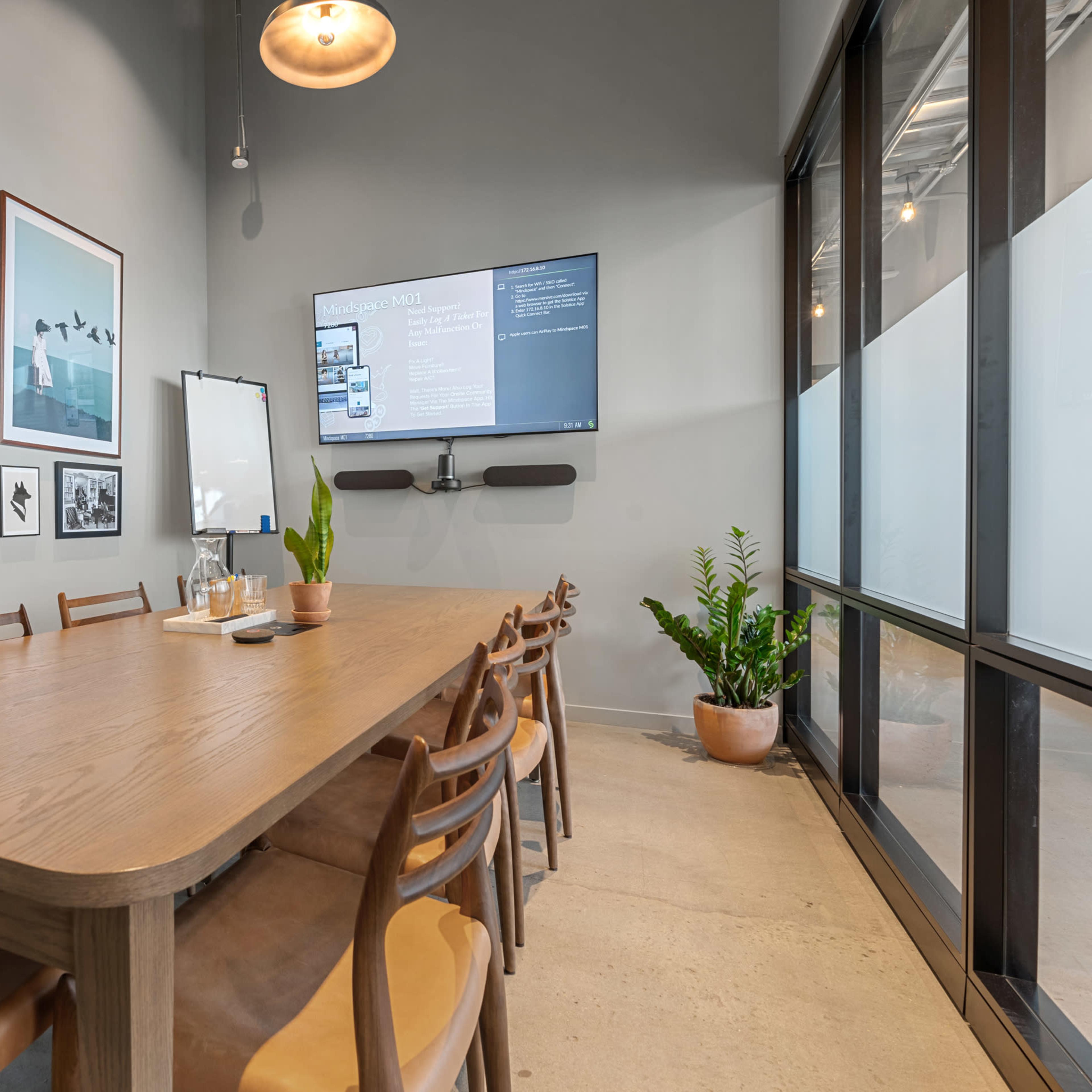 A conference room features a large wooden table surrounded by chairs, with framed photographs on the walls and a TV mounted on one side.