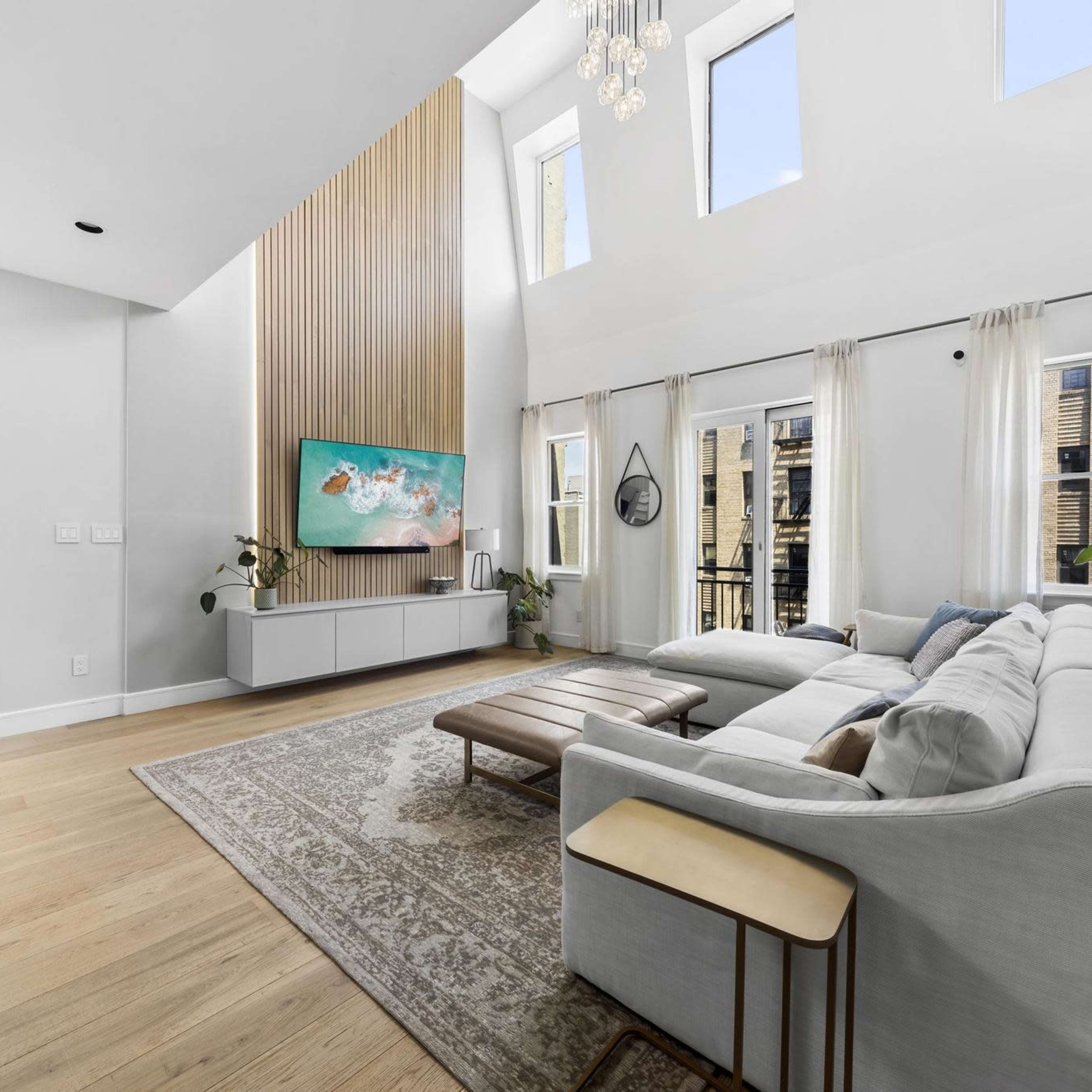 A spacious living room features a large sectional sofa, a wooden coffee table, and a mounted television against a tall, wood-paneled wall, with large windows allowing natural light.
