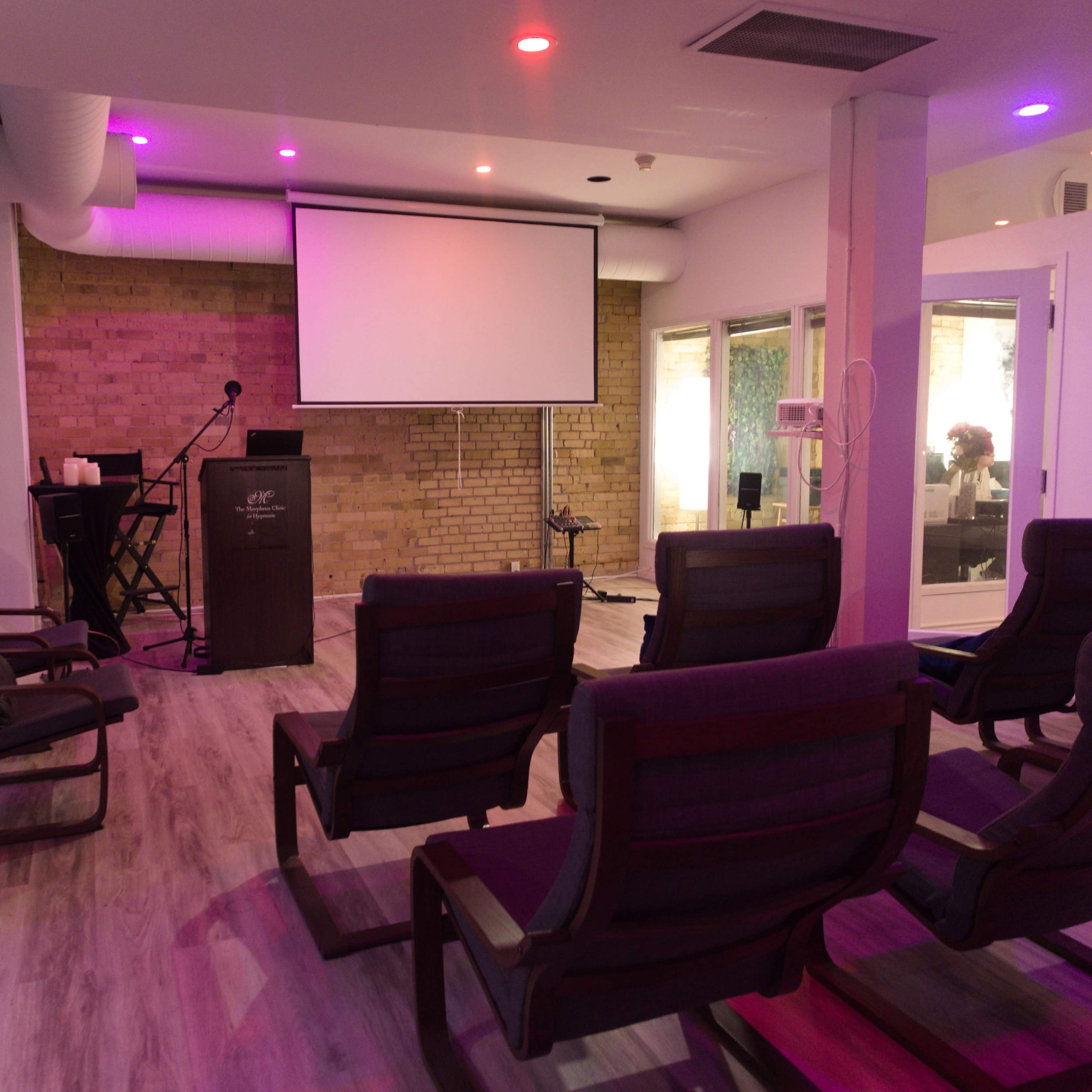 A small meeting room features several reclining chairs arranged facing a podium and a projector screen, with brick walls and ambient lighting.