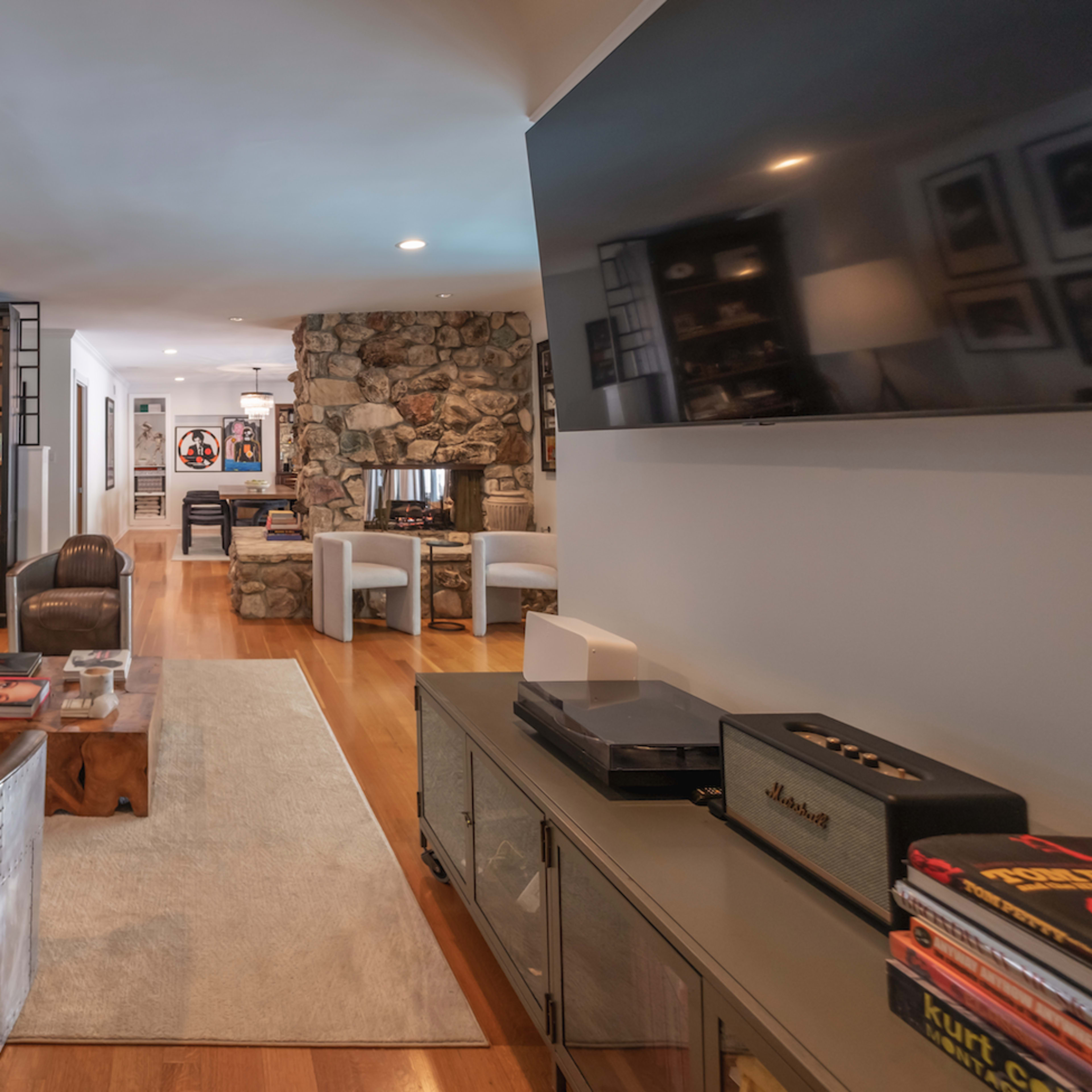 A modern living room featuring a leather sofa, a stone fireplace, a wall of framed photographs, and a TV mounted on the wall.