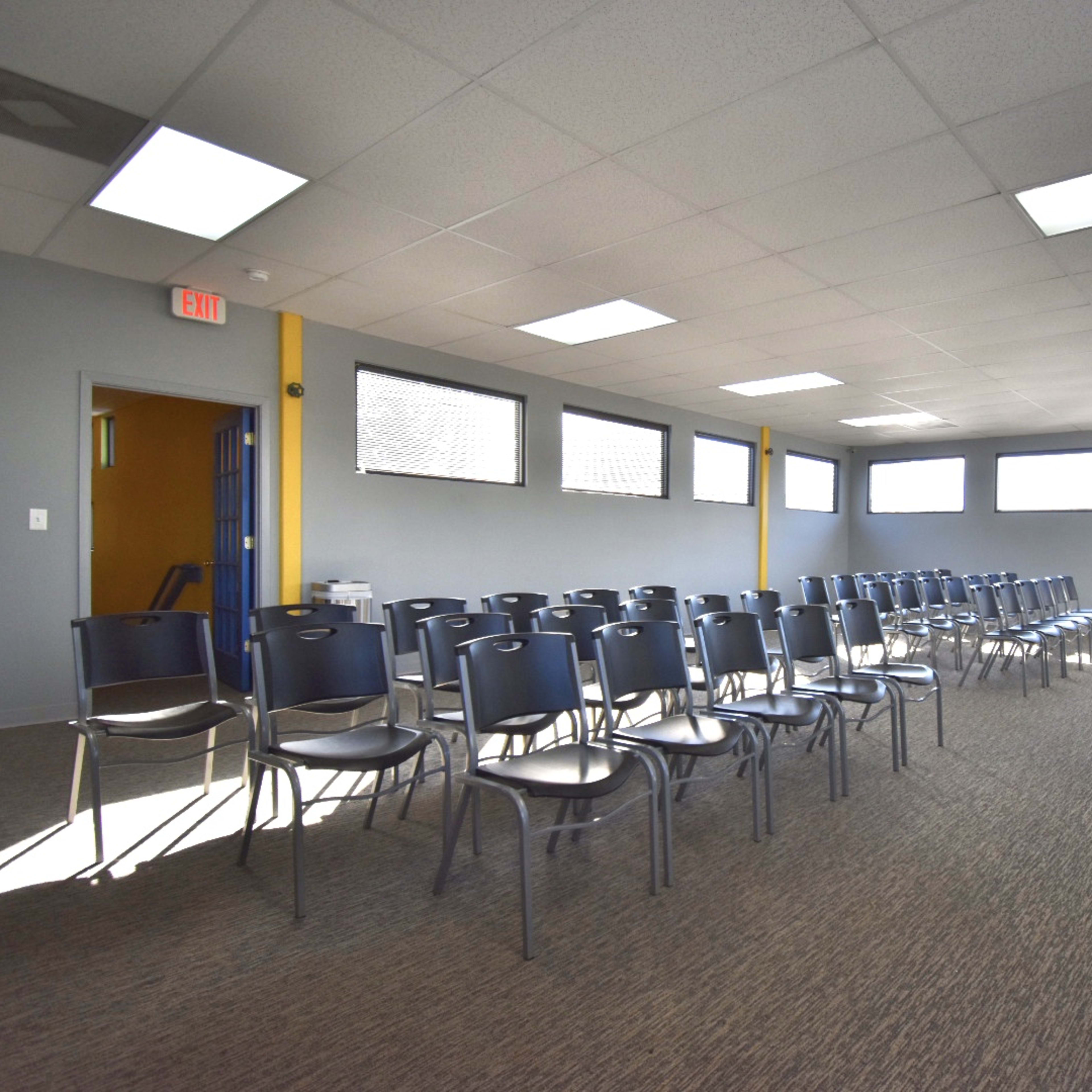 A row of chairs is arranged in a meeting room with a black table at the front and windows allowing natural light.