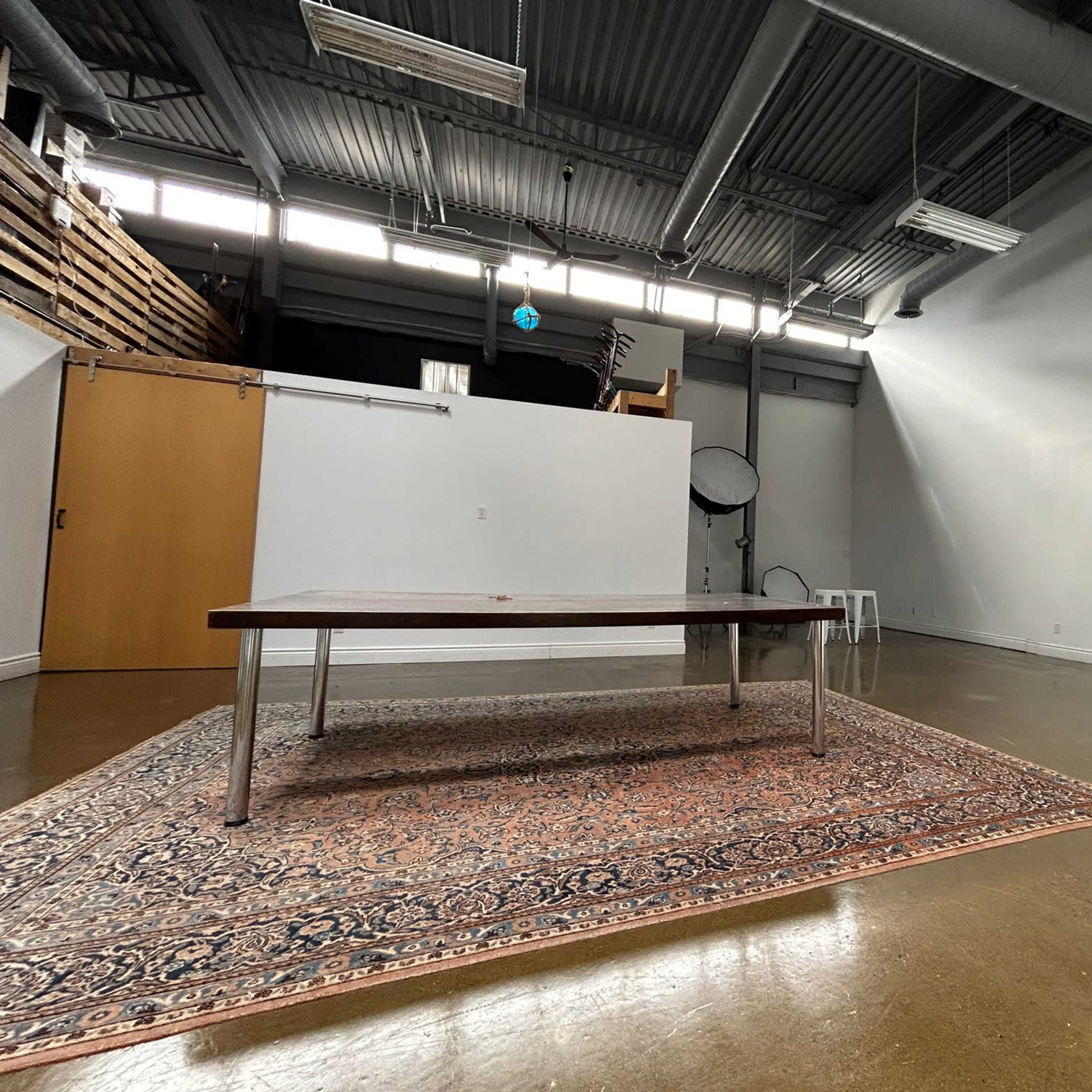 An empty room features a wooden table on a rug, with a backdrop visible in the distance.