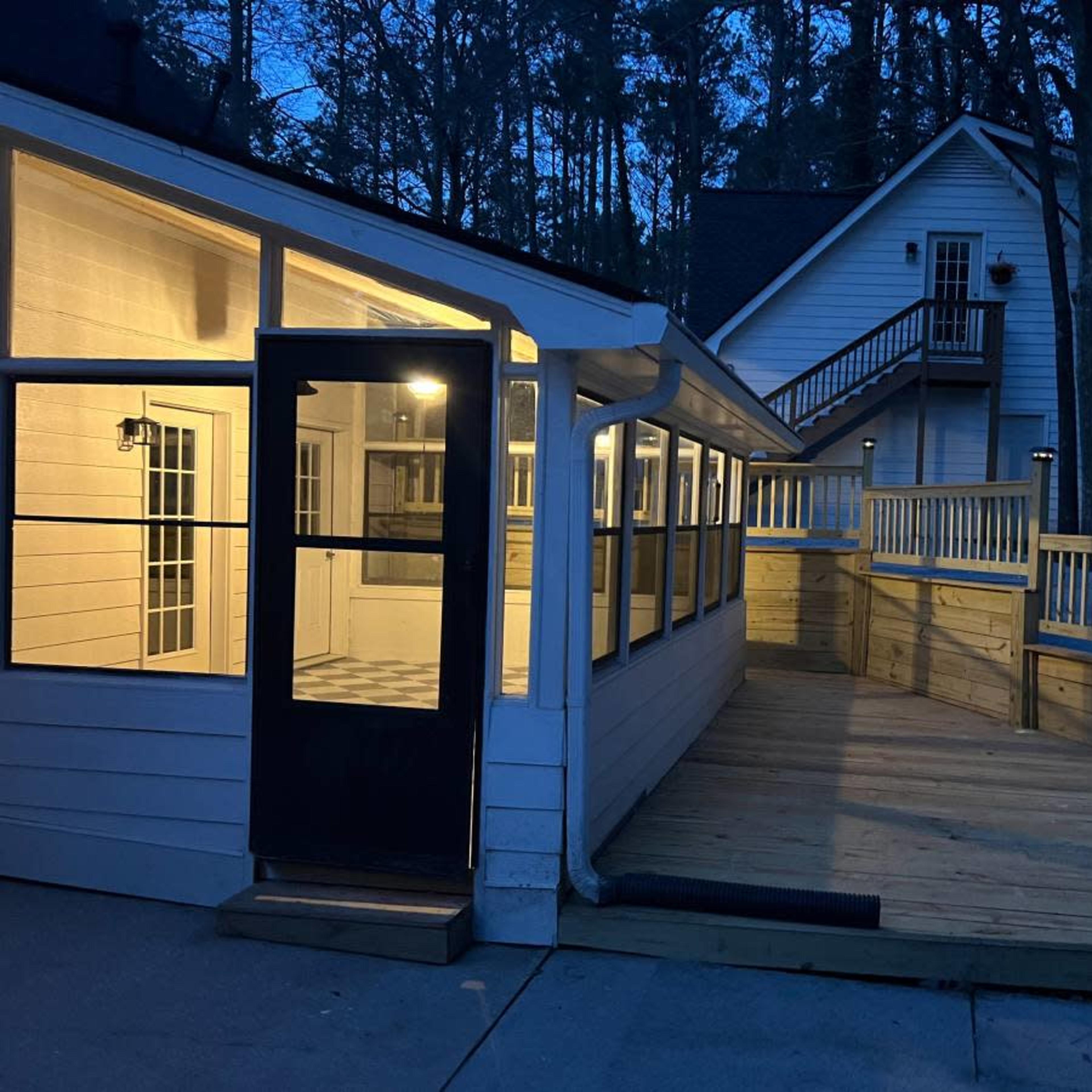 A wooden deck leads to a bright, enclosed sunroom attached to a house surrounded by trees at dusk.