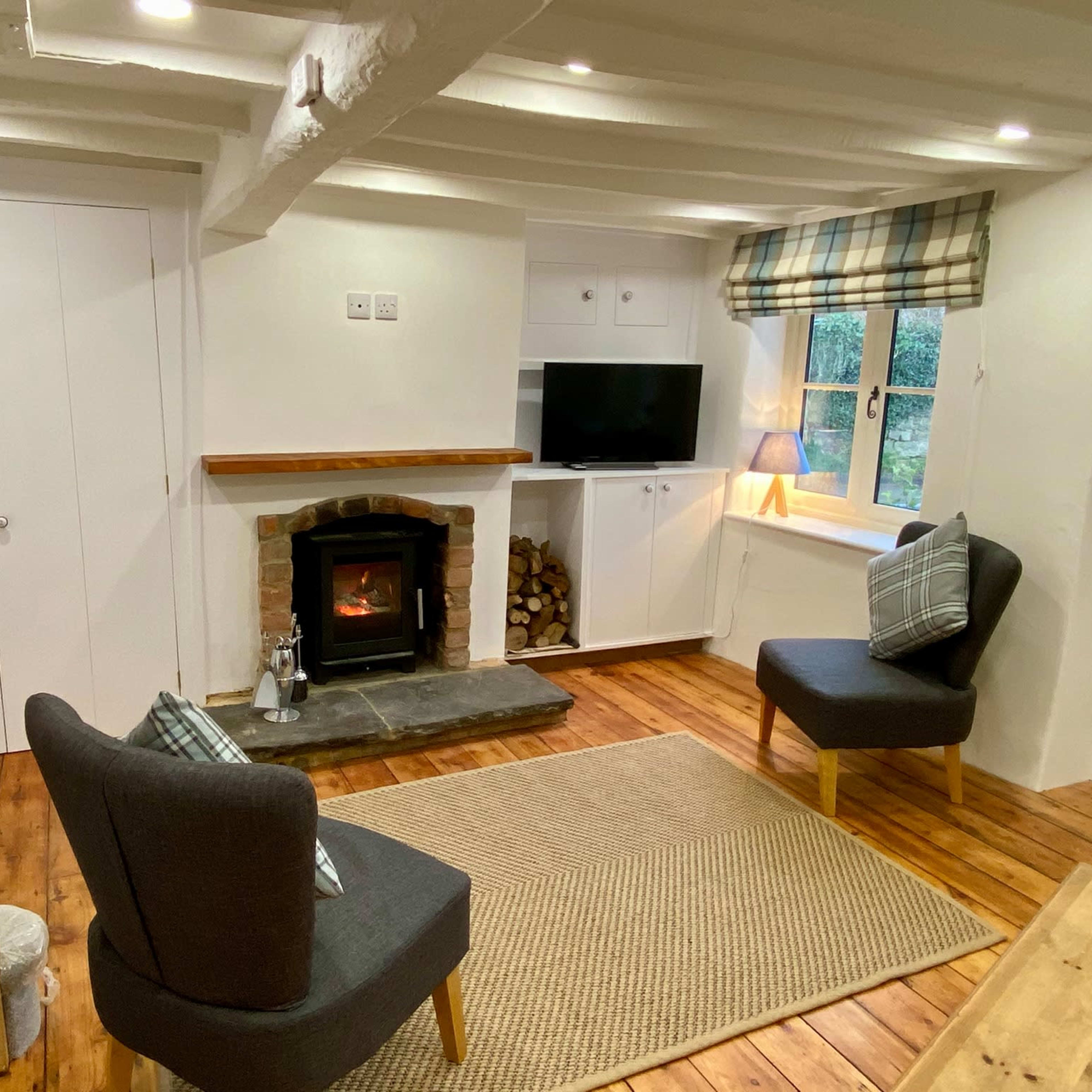 A cozy living room with two chairs facing a stone fireplace, a television mounted on the wall, and a window dressed in plaid curtains.