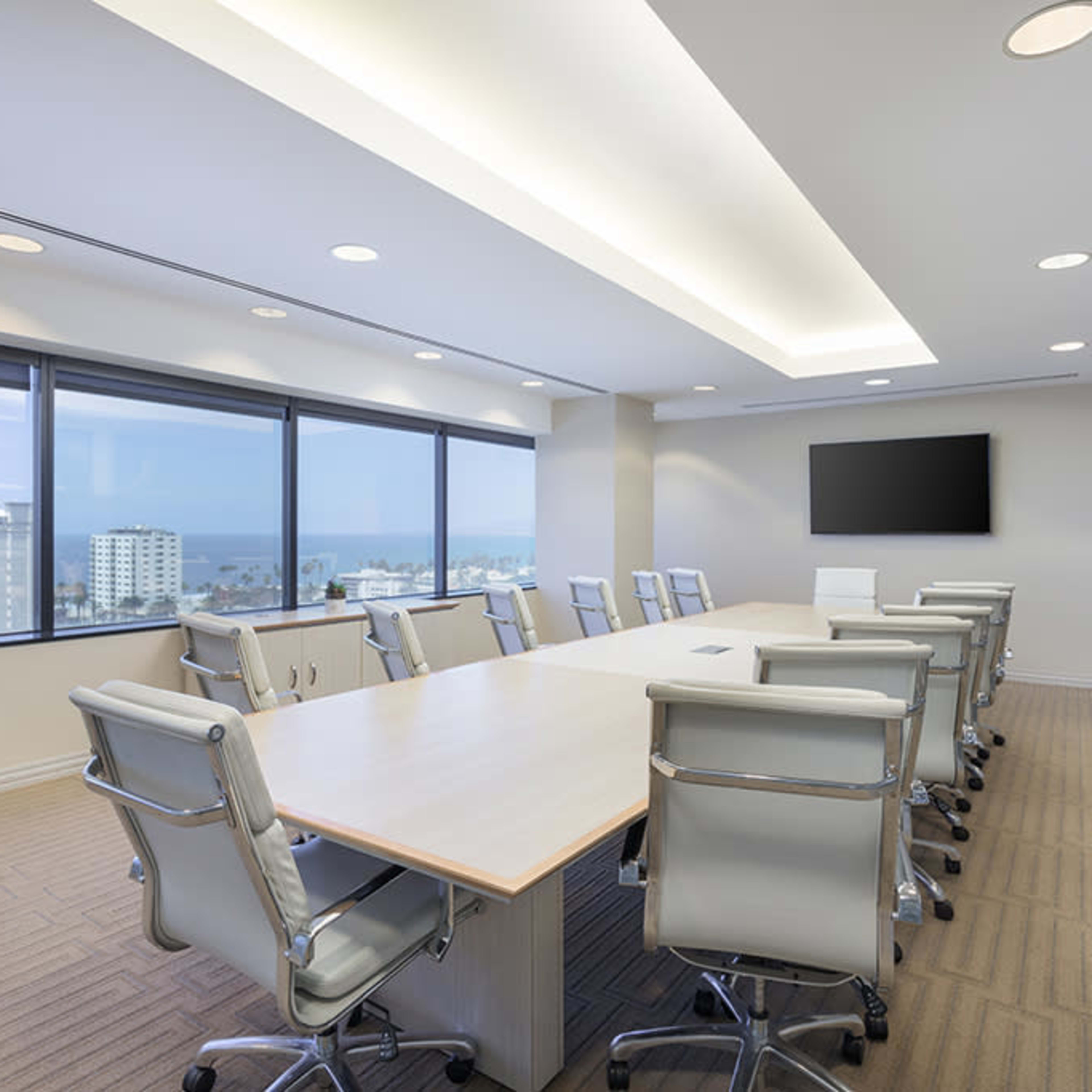 A modern conference room features a long table surrounded by ergonomic chairs, with large windows offering views of a cityscape and ocean.