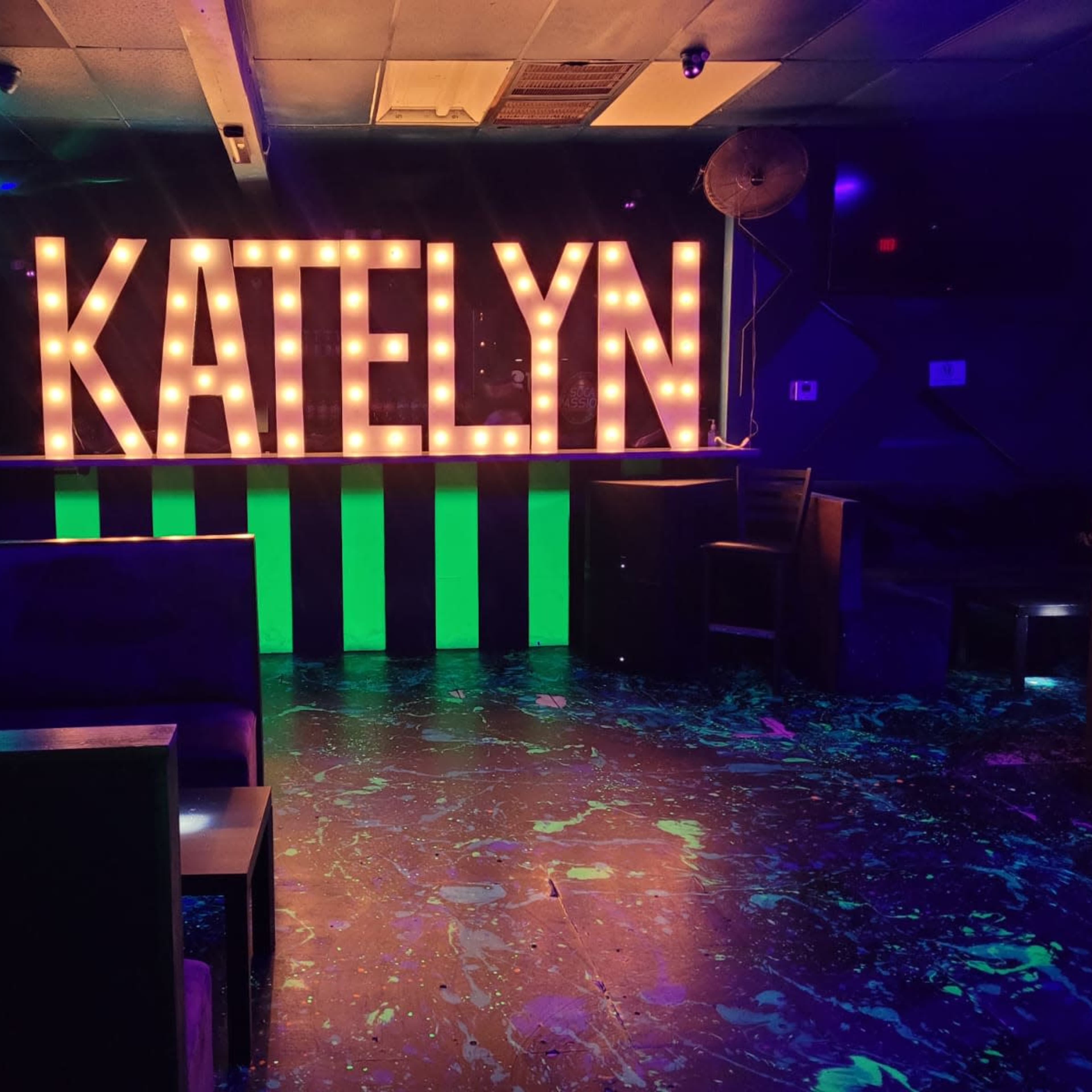 A dimly lit venue with a large illuminated sign spelling "KATELYN" and colorful decorations, including a green striped bar and dark seating areas.